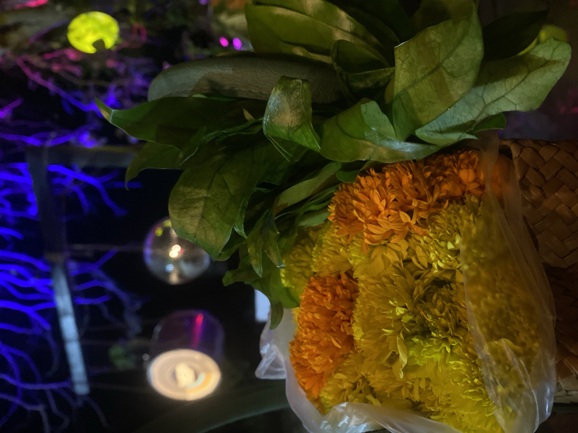 The image presents a floral arrangement, likely photographed in Baan Don Sai, Thailand, given the request's context. A bouquet of orange and yellow marigolds, accompanied by green leaves, is partially contained within a clear plastic bag. The flowers and leaves are placed in a woven basket.
In the blurred background, there is a light source that is greenish-yellow. There are also multiple decorative lights, including a few that appear to be silver or reflective, and another that is a warm white light. A blue light is also visible in the background, forming a vertical linear shape. The setting appears to be indoors or under artificial lighting conditions.
