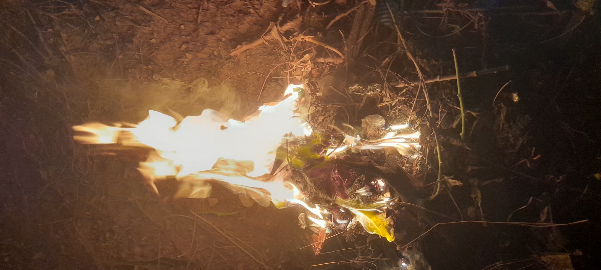 A fire is burning in a pile of dry leaves, branches, and other flammable debris in Buketteukuh, Indonesia. The flames are bright yellow and white, with smoke rising from the edges. The surrounding area is dark, with only the light from the fire illuminating the ground. The ground appears to be soil and leaf litter.