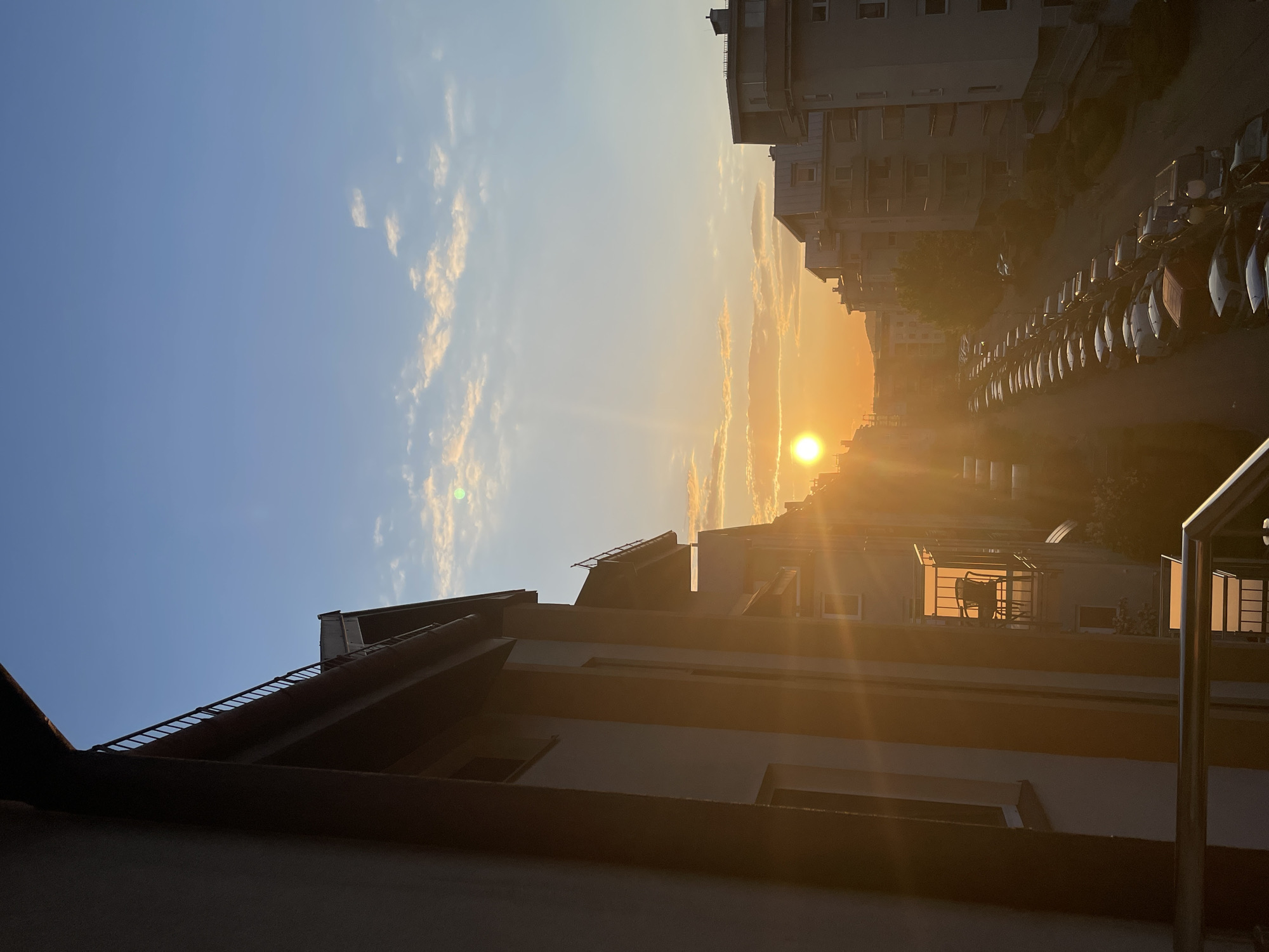 The image shows a sunset in a city. The sun is setting in the distance, obscured by buildings. To the left, the sky is blue with a few white clouds. To the right, a row of parked cars lines the street next to the buildings. A building with an angled roof is prominent in the foreground. The location is unknown, but it appears to be a residential area.