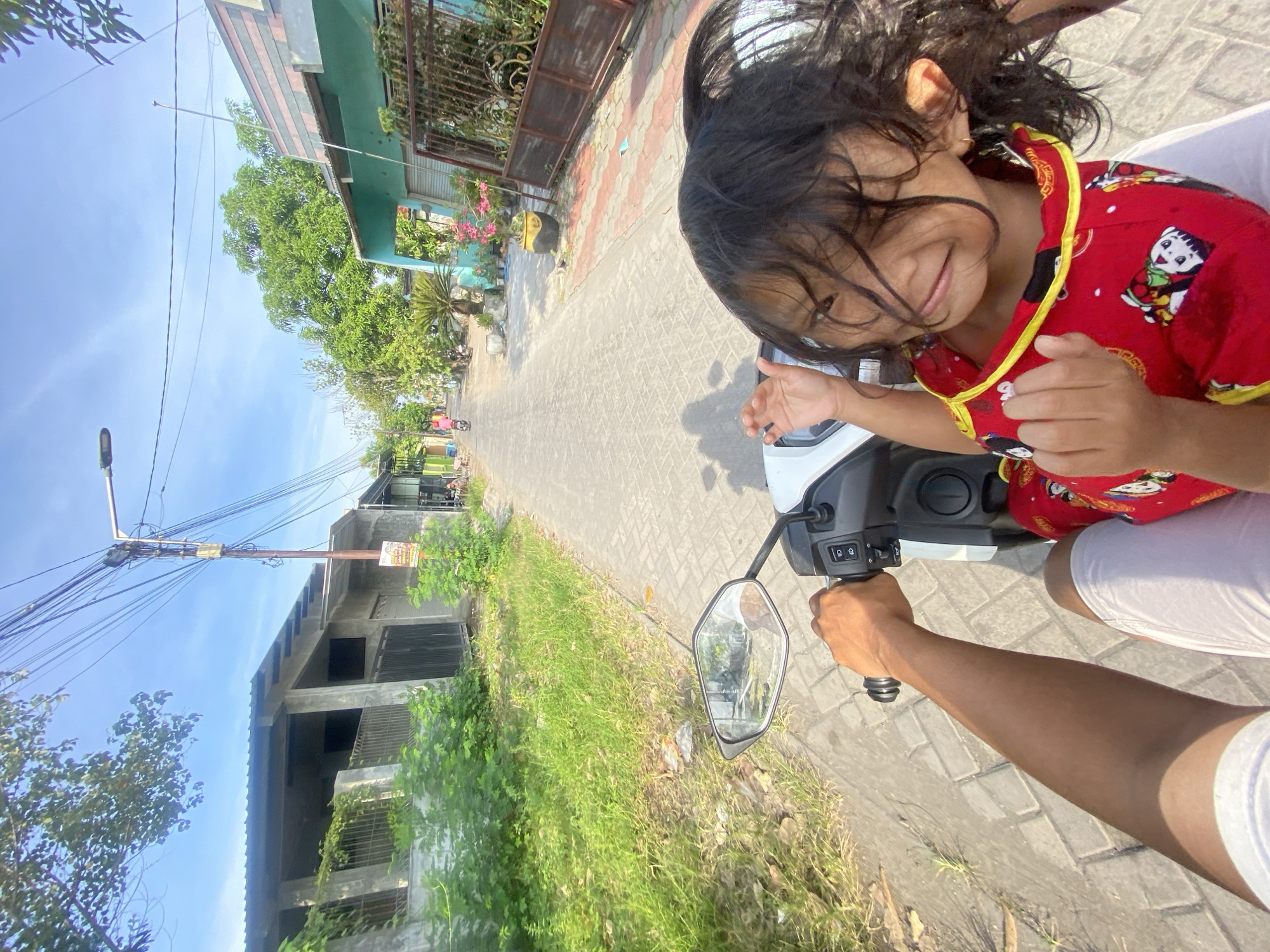 A medium-angle, eye-level shot captures a person driving a scooter on a paved road with a young child. The child, wearing a red, patterned top with yellow trim, is positioned in front of the driver. The driver's right arm is visible, holding the handlebar of the white scooter. A side mirror of the scooter is in view.
The road is paved with gray bricks. On either side of the road, there is green grass. A building with exposed concrete construction can be seen to the left. A utility pole with numerous wires extends diagonally across the top left corner of the image. A house with a green roof is seen on the right side of the image with a brown metal gate. The sky is light blue with faint clouds.
Based on the surroundings, the location could be Gelam, Indonesia, identified by the distinctive building styles and environment.