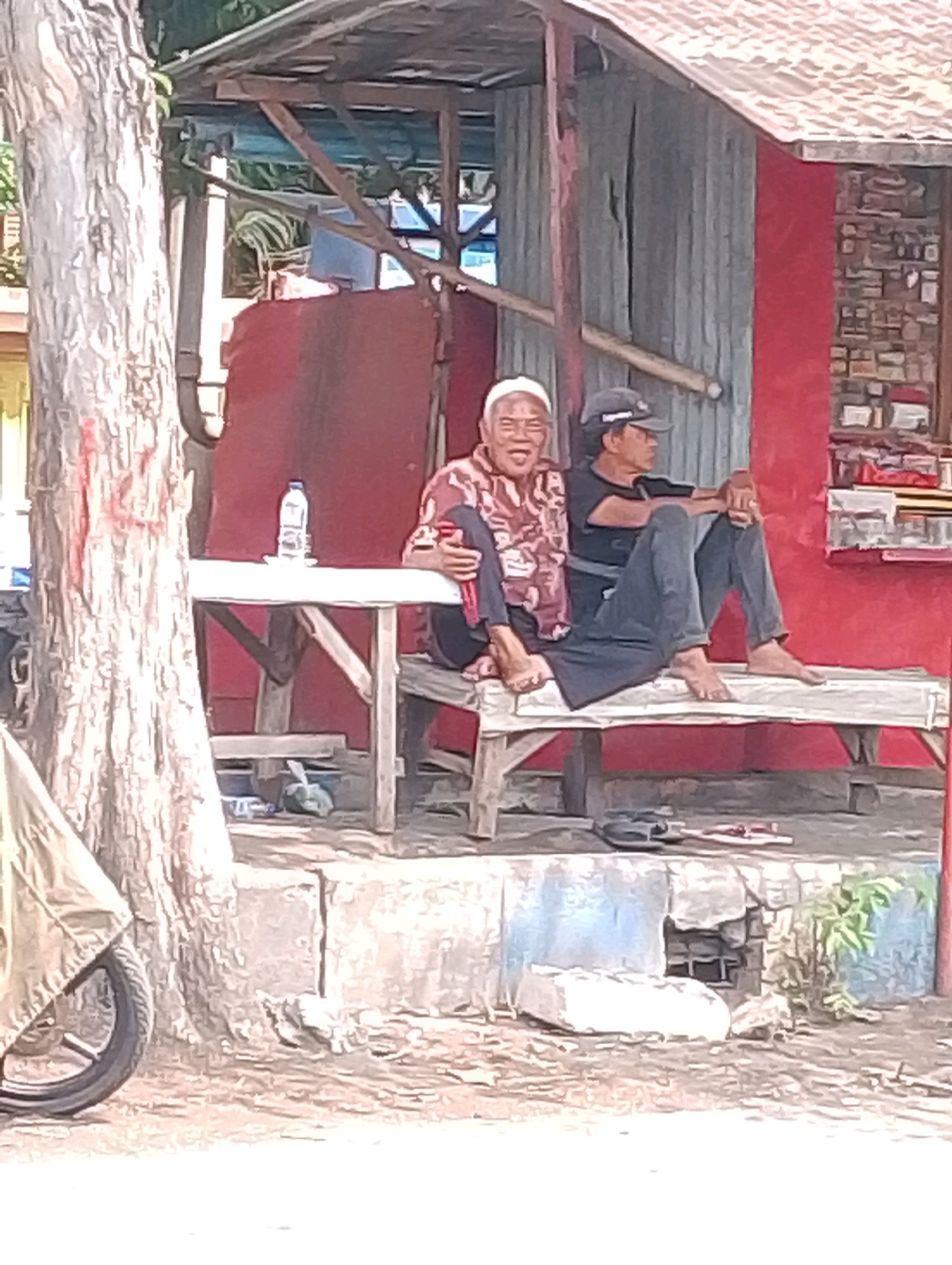 Two men are sitting on a wooden bench in front of a red building with a corrugated metal roof. The man on the left is wearing a white cap and a patterned shirt, and he is smiling broadly. The man on the right is wearing a dark baseball cap and a dark t-shirt, and he is looking to his right. Both men are barefoot. A white table with a plastic water bottle on it is to the left of the bench, near a tree trunk. The ground in front of the building is covered in dirt and gravel, with some scattered debris. A motorcycle wheel is visible on the far left of the image. The scene appears to be an outdoor seating area, possibly at a small shop or food stall in Probolinggo, Indonesia.