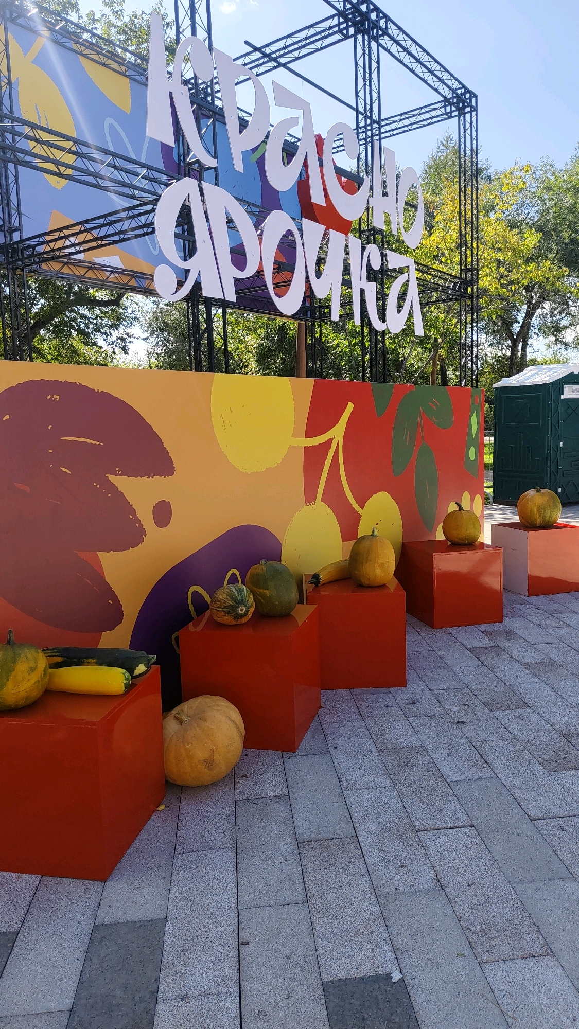 The image depicts an outdoor scene, likely a public event or market in Krasnoyarsk, Russia. In the foreground, several pumpkins and gourds are arranged on bright orange cubic pedestals. A large, colorful mural featuring abstract shapes and fruit-like designs serves as a backdrop. Above the mural, large white letters spell out "КРАСНАЯ ЯРОЧКА" (Krasnaya Yarochka), a name or slogan, which are mounted on a black metal scaffolding structure. Trees with green foliage are visible behind the scaffolding, suggesting a park or outdoor venue. To the far right, a green portable toilet is partially visible. The ground is paved with light-colored rectangular stones. The scene appears to be set up for a harvest festival or a similar autumnal celebration. There are no people visible in the image.