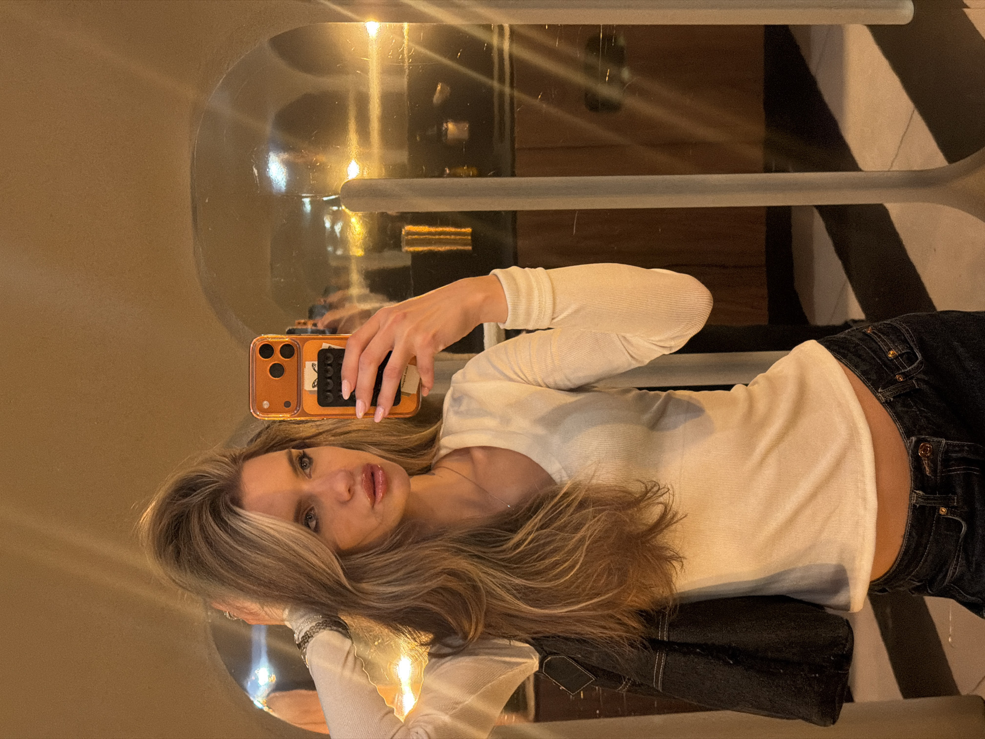A young woman with long, blonde hair is taking a selfie in a mirror. She is wearing a white long-sleeved shirt and dark jeans. A black jacket is visible behind her. The mirror is part of a semi-circular structure with a chrome frame. In the reflection, a striped wall and a shelf or counter are visible. The location is Berlin, Germany, likely indoors at a bar or restaurant. The woman is holding an orange iPhone. The time of day is likely evening or night due to the indoor lighting.