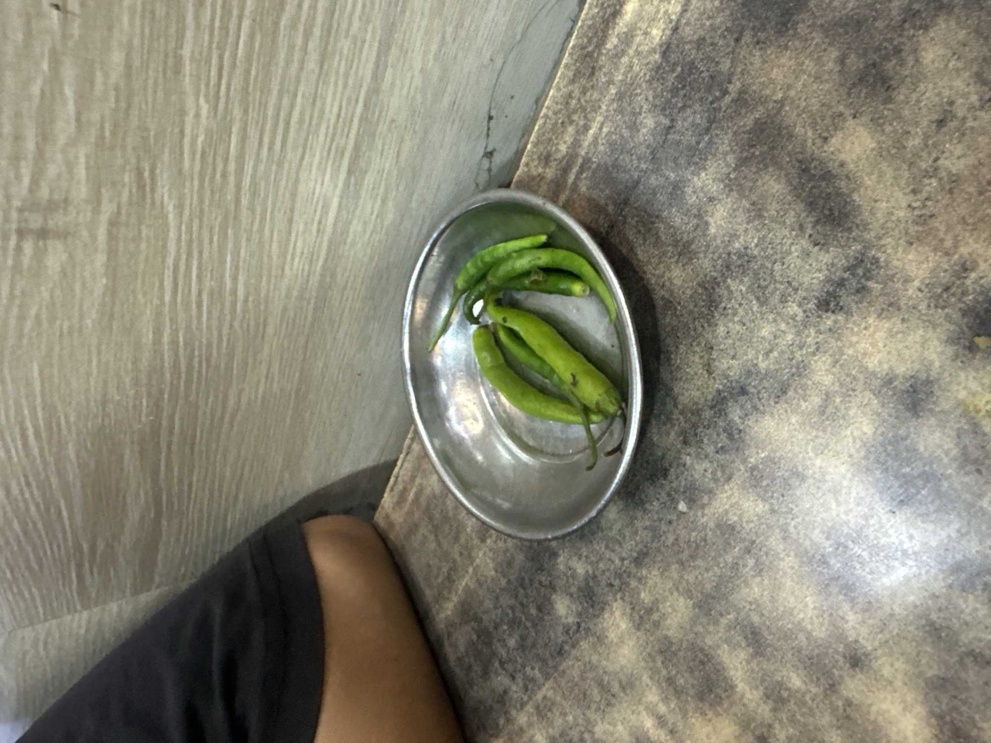 A shallow, tarnished metal bowl contains a cluster of six bright green chili peppers on a textured, dark brown surface. The bowl is positioned on the left side of the image. To the left of the bowl, a person's arm, clad in a black t-shirt, is visible, resting on the surface. The background consists of a light-colored, wood-grained wall. The scene appears to be indoors, possibly in a restaurant or dining area, given the presentation of the chili peppers.