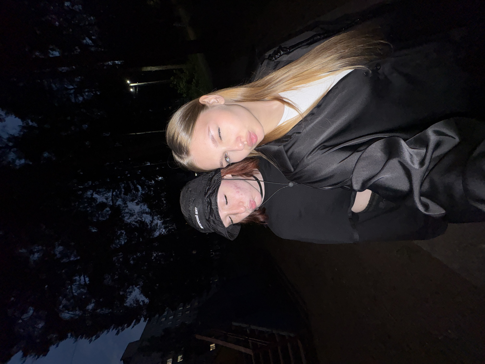 In a nighttime outdoor scene on the outskirts of Chaikovsky, Russia, two young women are captured in a close-up, low-angle shot. The woman on the left, wearing a black bucket hat with white lettering, faces away from the camera with her head tilted down and lips pursed. She has light skin with visible acne. The woman on the right, with long blonde hair, winks at the camera and also pouts her lips. She is wearing a white top and a shiny black jacket. Behind the women, the environment is dark and appears to be a park or wooded area, with trees silhouetted against a twilight sky. A tall, dark building with illuminated windows is visible to the far left, and a streetlamp can be seen in the background on the right. The ground in the foreground is dark and indistinct.