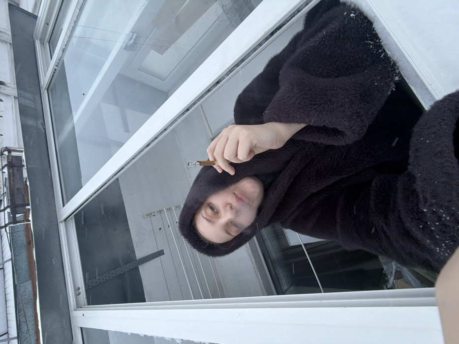 The image, when viewed upright, captures a close-up perspective of a person looking directly at the camera, seemingly from a window or balcony on a cold day. The central figure is a person, likely female, with fair skin and light-colored eyes, wearing a dark, possibly black or very dark brown, hooded coat or robe made of a fuzzy or furry material. Small white specks, consistent with snowflakes or ice crystals, are visible on her coat, particularly on the sleeves and shoulder, and also on the white window frame.
Her right hand is raised, holding a lit cigarette or a similar rolled smoking item between her fingers, with a glowing ash tip and a filter clearly visible. Her facial expression is neutral, appearing calm and perhaps a little contemplative.
The immediate foreground is dominated by a clean white window frame, which divides the view into several panes. Through the window, in the background, elements of an outdoor building structure are visible, including a grey wall on the left with some industrial-looking metal pipes or fixtures attached. The overall lighting is soft and diffuse, suggesting an overcast sky typical of winter. The presence of snow or frost indicates cold weather. The scene conveys a quiet, possibly solitary moment. No text is visible in the image.