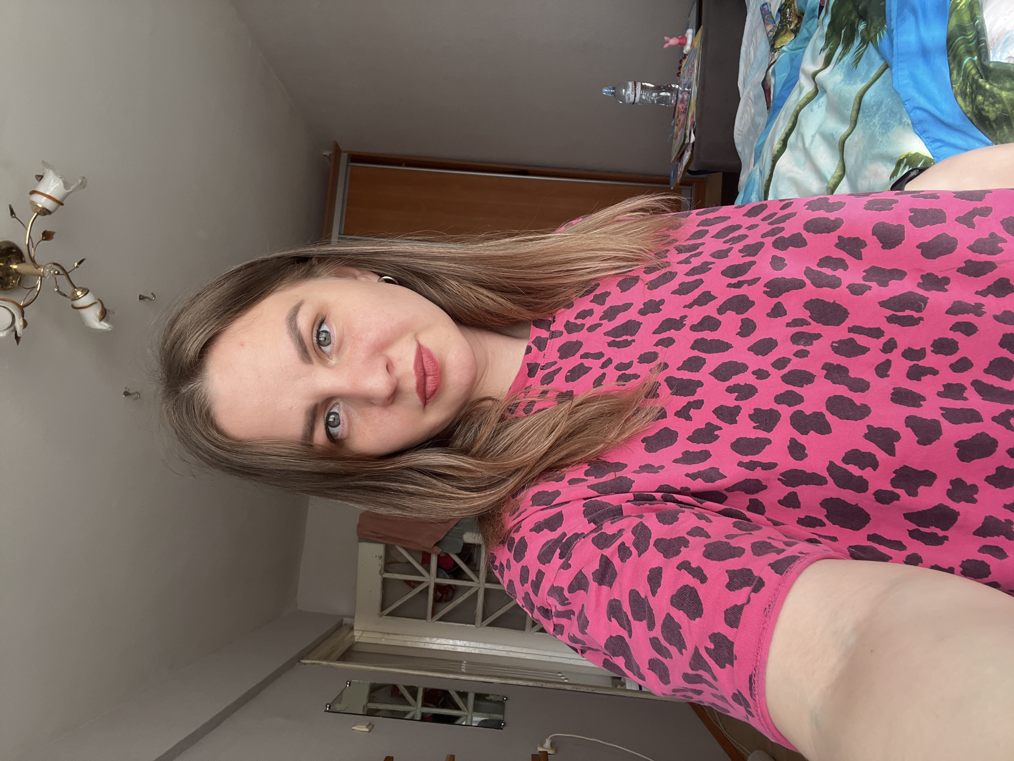A selfie captures a young woman, appearing to be lying down, looking directly into the camera. The image is rotated 90 degrees clockwise. She has long, light brown hair and is wearing a vibrant pink top with a dark leopard print. Her expression is neutral to slightly smiling, and she wears subtle makeup, including a pinkish-red lipstick. A small hoop earring is visible on her left ear.
The setting is an indoor space, likely a bedroom, in Votkinsk, Russia. The background reveals various elements of the room. To the upper left, an ornate, gold-colored ceiling light fixture with white, tulip-shaped shades is mounted on a light grey ceiling. Behind the woman, a wooden wardrobe or cabinet is partially visible. Further in the background, a bed is made with a blue and green patterned duvet or blanket, possibly featuring a tropical or floral design. On a surface near the bed, a plastic water bottle with "Chistaya Voda" written on it is present, alongside a small white figurine and some papers. Towards the bottom of the frame, a small rectangular mirror reflects a part of the room, positioned below what appears to be a white latticed or framed structure. The lighting suggests it is daytime.