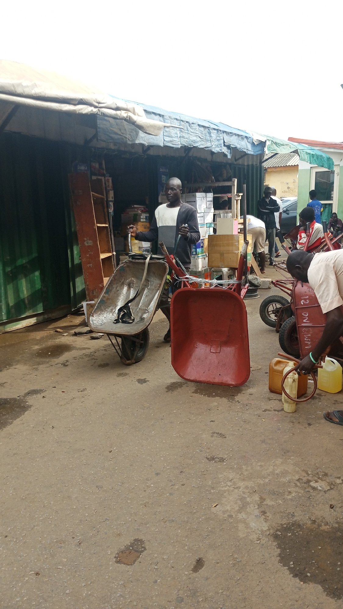 An outdoor scene in Abuja, Nigeria, depicts a marketplace or roadside stall. In the foreground, two wheelbarrows are visible, one a rusty metal one with a black strap and the other a red one. Several plastic containers, some yellow and some transparent, are also present on the ground, with one being filled from a dispenser. A man wearing a grey and white sweater is standing next to the metal wheelbarrow, holding its handles. Behind him, there are shelves stacked with goods, possibly in boxes. Further back, other individuals are seen standing and interacting, and a car is parked in the background. The area appears to be paved with concrete, and there are some water or liquid stains on the ground. The overall setting suggests a commercial area, possibly involved in the sale of goods or supplies.