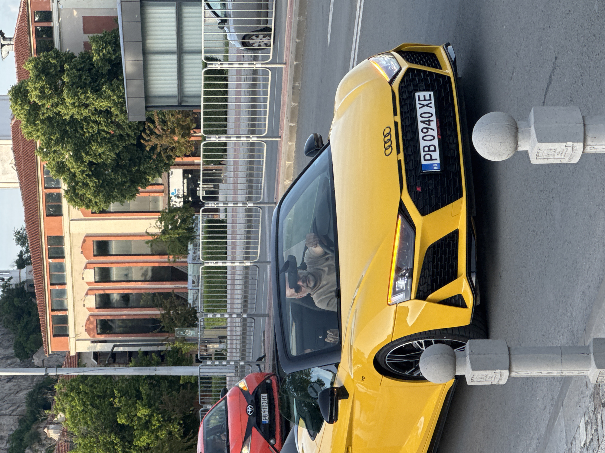 A medium shot captures a yellow Audi RS car with the license plate "PB 0940 XE", positioned on a street in Plovdiv, Bulgaria. A driver is visible behind the wheel, wearing sunglasses and a beige shirt. A red Toyota car with license plate "PB 9103 HT" is parked nearby. The street is lined with metal barriers. A multi-story building with peach-colored walls and dark-framed windows, along with lush green trees, are in the background. A concrete post is in the foreground, close to the Audi.