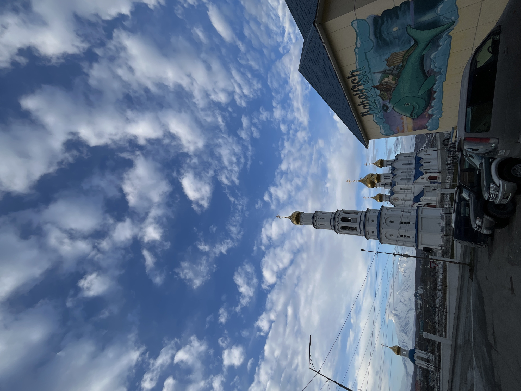 The image shows a view in Petropavlovsk-Kamchatsky, Russia, dominated by a bright, blue sky filled with scattered clouds. A white Orthodox church with blue and gold domes and a tall bell tower stands prominently in the midground. To the right of the church are parked cars. In the background, snow-capped mountains are visible. A building with a mural of a whale is partially visible at the top right. A street lamp and electrical wires are present in the foreground.