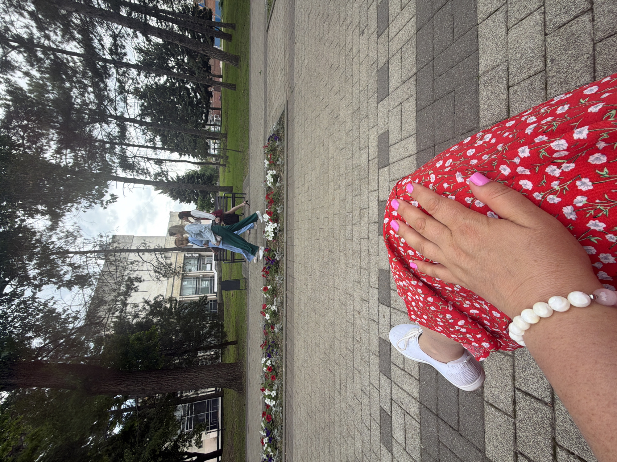 The image shows a person wearing a red dress with white floral print and white shoes, sitting on a gray and white brick pathway. The person is also wearing a bracelet made of white beads. Their fingernails are painted pink. In the background, there is a line of trees along a grassy area with a flowerbed. Further back, there is a building and a group of three people sitting on a bench. The location appears to be in a park.
The image also contains enough detail to suggest that the city is Leningradskaya, Russia, as the style of architecture in the background matches buildings commonly found in that city.