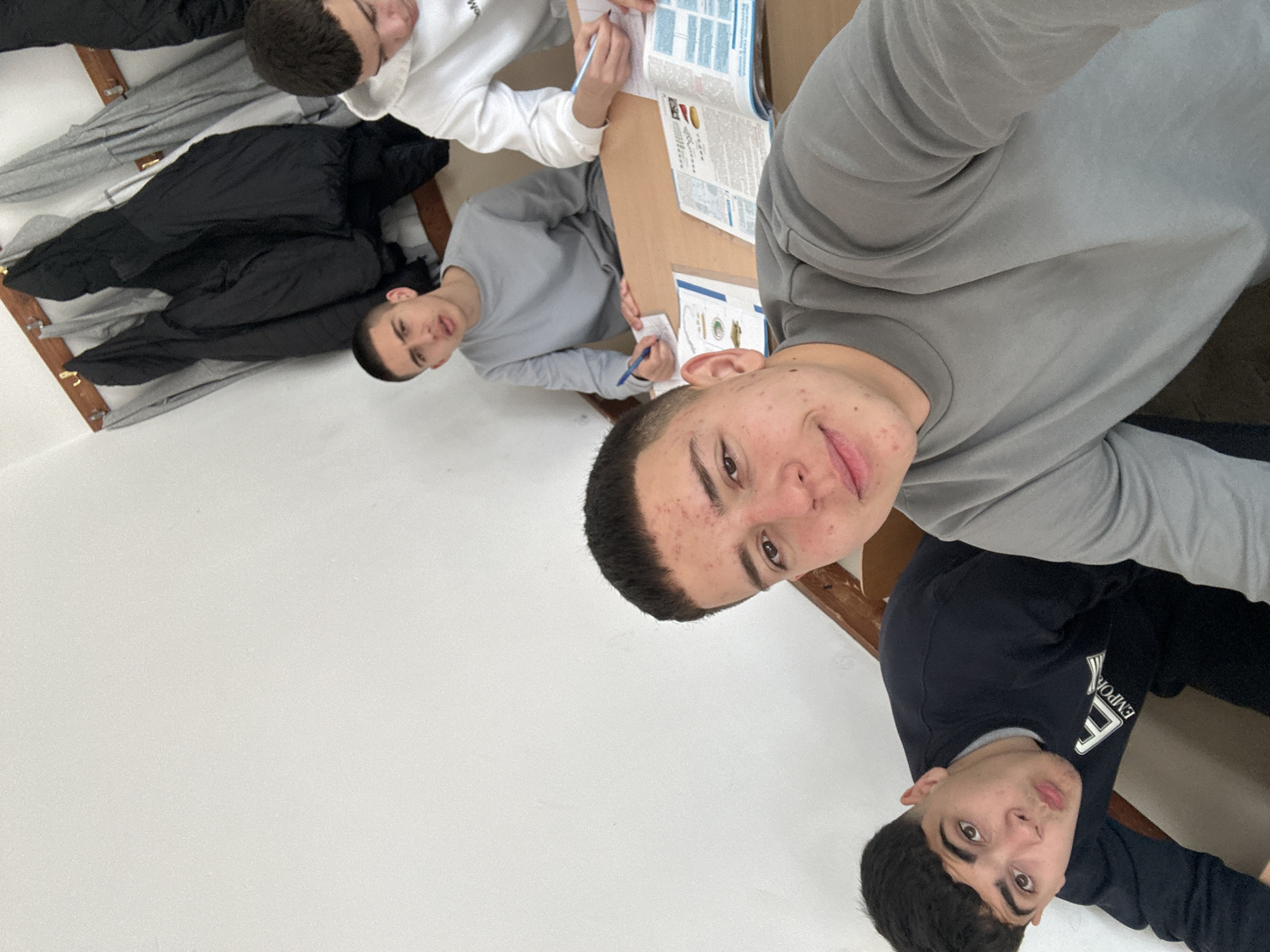 The image, taken in Zlatograd, Bulgaria, depicts an indoor scene with four young men, captured at an angled perspective as if from a selfie.
In the prominent foreground, a young man with short dark hair and light acne is looking directly at the camera with a slight smile, wearing a grey long-sleeved shirt. To his right, another young man is partially visible, wearing a black long-sleeved shirt with a white "EMPORIO" logo, also looking towards the camera.
Further into the frame, two other young men are seated at a light-colored wooden table, engaged in studying or work. One, in a grey long-sleeved shirt, is writing with a blue pen on a notepad. Behind him, another individual in a white hoodie with a visible "W" on it is also focused on an open book or papers spread across the table.
The setting appears to be a brightly lit room with white walls, possibly a classroom or study area. A black jacket is draped over a piece of furniture in the background. The overall activity suggests a group study session.