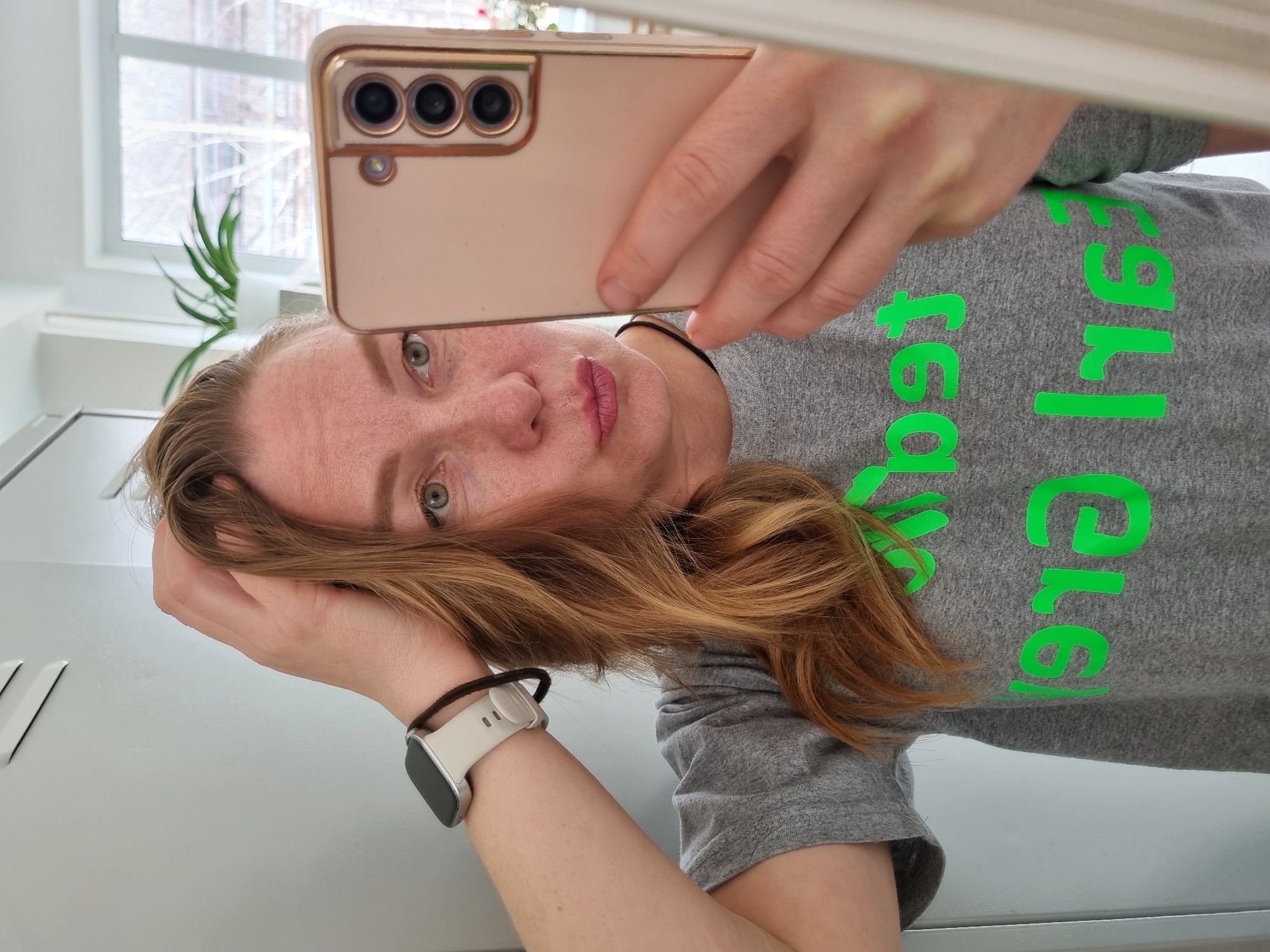 A woman is taking a selfie in what appears to be an indoor setting. She is holding a smartphone up to capture the image, the phone's camera array is visible. The woman has fair skin, reddish-brown hair, and is wearing a gray t-shirt with green writing on it. She is also wearing a smart watch on her left wrist. In the background, there is a window and a plant. The setting is minimalistic, suggesting a modern interior. There is no indication of Kemul, Russia in the image.