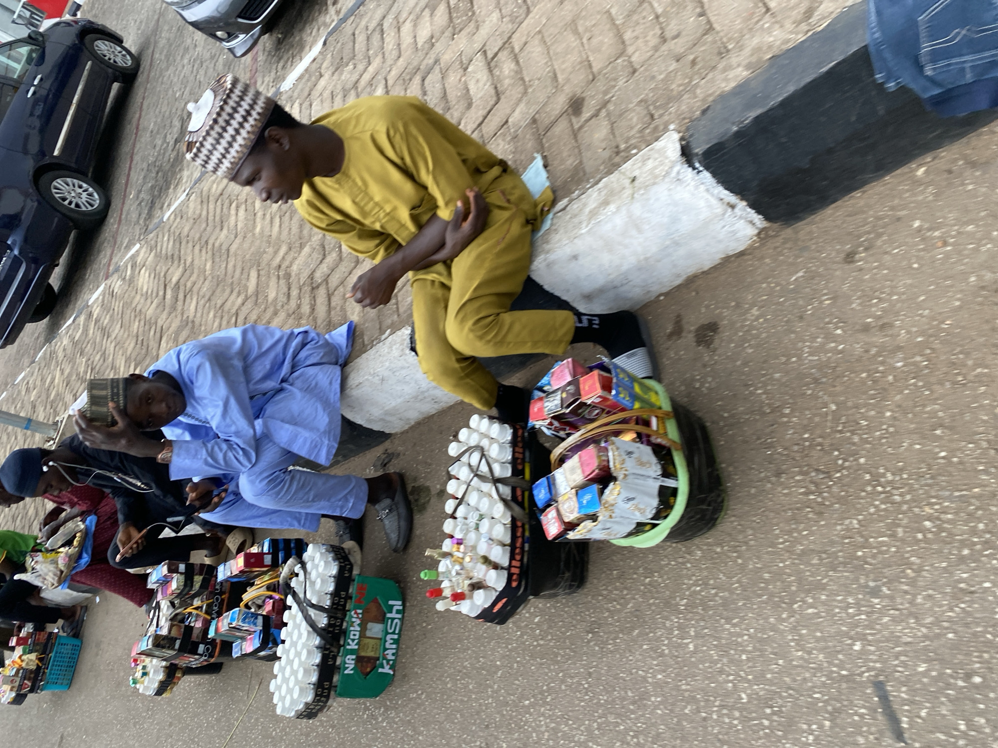 The scene depicts a street in Jos, Nigeria, with several individuals seated on a curb. A man in a yellow robe and checkered hat is sitting next to a black curb. Adjacent to him, a man in a blue outfit sits holding a box to his head. Both are near displays of small goods. Further along, another man in a black hat and black attire is holding a phone. A man in a red top sits next to him. These individuals have displays of small goods, potentially for sale. The background features cars parked on the side of the road. The curb is painted in black and white, the street paved in grey stone and the overall setting suggests a commercial area.