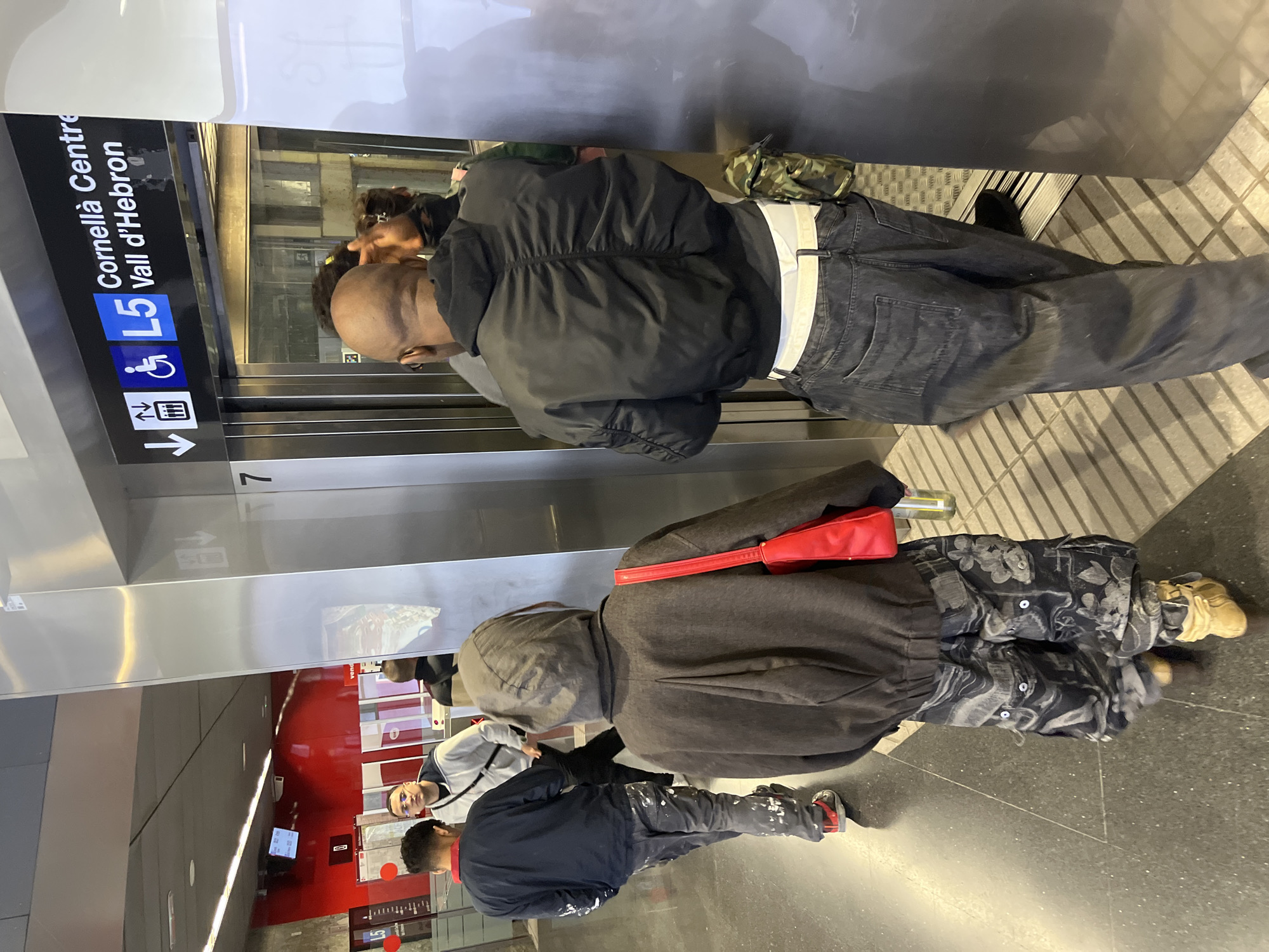 The image appears to be taken inside a metro station in Barcelona, Spain, based on the "L5" metro line sign indicating "Cornellà Centre - Vall d'Hebron." There are multiple individuals present. Two people are inside the elevator, another is standing by the elevator, and another two are standing in the main station area. The elevator is silver with a dark grey interior, with a sign indicating the accessible access, and the number 7 is visible. In the main station area, there is an area with a red wall and signs. The flooring of the station appears to be grey and polished.