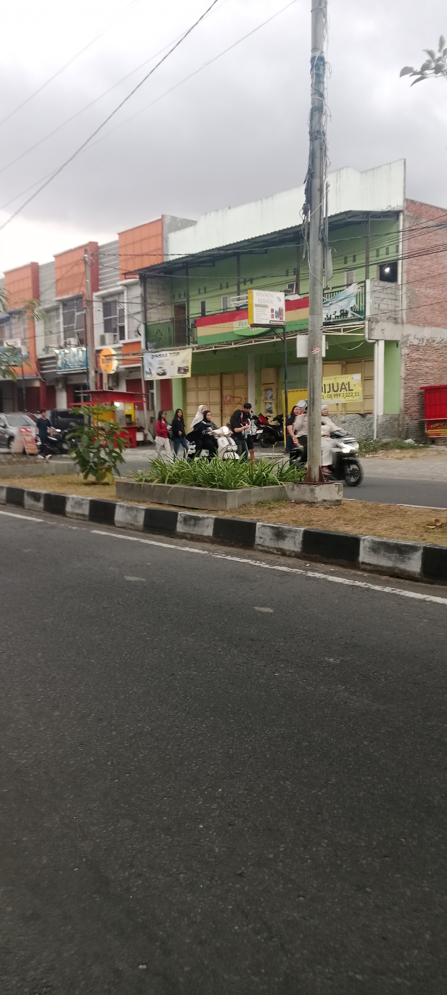 This is a full, slightly tilted, eye-level shot of a street scene in Ngemplak, Indonesia, under an overcast sky. The foreground is dominated by an asphalt road with a white dashed line. A grey concrete barrier with black and white stripes separates the road from a raised planter with sparse green foliage and dry grass. In the middle ground, a row of shops with multi-colored facades runs along the street. The buildings have multiple stories, some with balconies and air conditioning units visible. A tall, grey utility pole with wires and peeling insulation stands prominently near the center of the image. Several people are present, mostly on or around motorcycles. One group of women walks along the sidewalk. A man in a white shirt and dark pants is visible near a parked motorcycle. Two people are riding a white scooter in the middle of the street. Another person is riding a black motorcycle to the right of the scooter. A red cart or food stall is parked in front of one of the shops. Signs are visible on the buildings, including one with the words "DIJUAL" and a phone number. The overall impression is of a typical urban street environment in Indonesia.