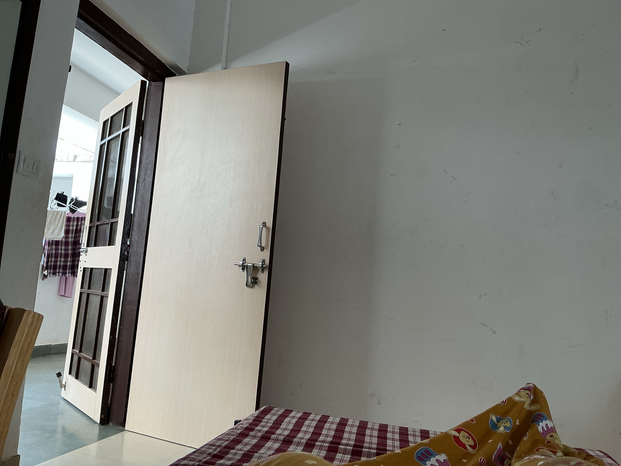 The image shows an interior scene, likely a bedroom, with an open door leading to another room or hallway. The room is sparsely furnished. A plain, light-colored door is a prominent feature, angled open and revealing a dark wooden door frame. On the door, there is a silver latch and handle. To the left of this door, another door is visible, partially open, with a glass panel and wooden frame. Laundry is hanging on a rack in the background, visible through the partially open doorway. The walls are plain and light-colored. In the foreground, a mattress with a red and white checkered cover is visible, partially covered by a yellow blanket with cartoon animal prints. The floor appears to be tiled in a light gray color. The lighting suggests an indoor environment, possibly during daylight hours due to the light coming from the open doorway. The scene appears to be in a residential setting, consistent with a room in Vanasthali, India. There are no people visible in the image.