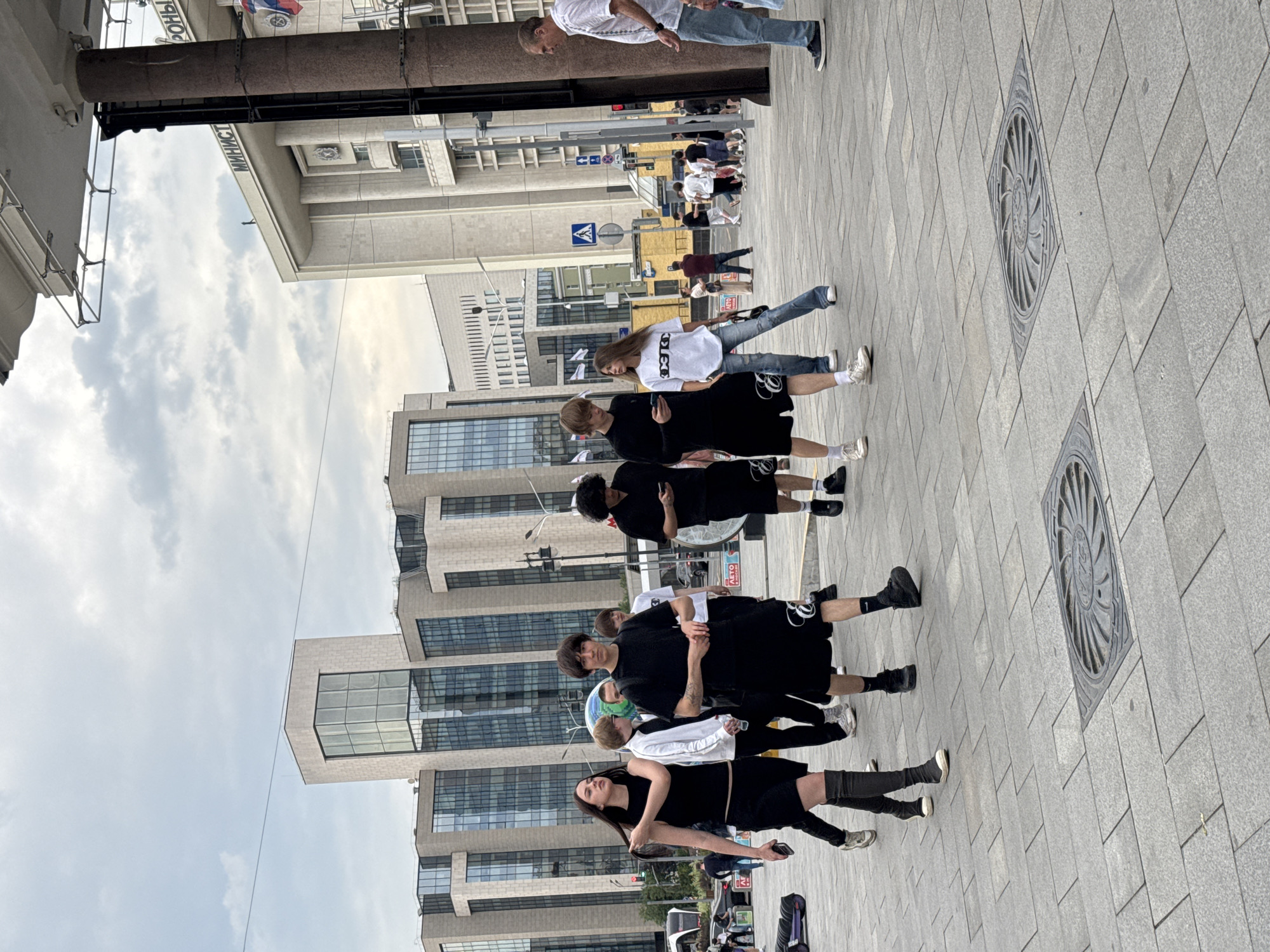 The image depicts a group of young people walking on a wide, paved sidewalk in an urban environment, likely in Moscow, Russia, given the architecture and signage. In the foreground, a group of five individuals, all dressed in dark, casual attire, are walking towards the viewer's left. A woman with long dark hair in the front is wearing a black sleeveless top and a black skirt, with tall grey boots. To her right, a person in a black t-shirt and shorts is visible. Behind them, three more individuals in dark clothing are walking. The background features modern, tall buildings with glass facades and some older, grander architecture with visible text, possibly governmental or institutional, including the word "МИНИСТ" which suggests "Ministry" in Russian. A tall, dark brown column, likely part of a building or structure, stands prominently on the right side of the frame. The sky is overcast with clouds. Several manhole covers are embedded in the pavement. The overall scene suggests a busy, public space in a city.
