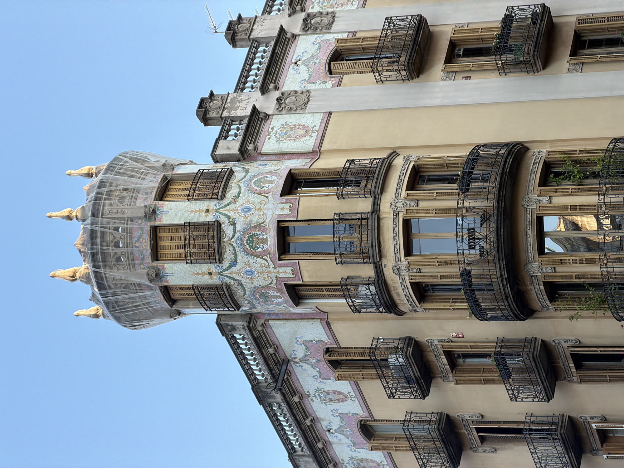 The image captures a close-up, low-angle view of a historic building in Barcelona, Spain, bathed in the warm light of either sunrise or sunset. The building, characterized by its ornate Art Nouveau architecture, features a prominent round turret topped with a netting structure. The turret is adorned with colorful mosaic tiles depicting floral motifs and baskets, interspersed with windows featuring wooden shutters. Several balconies with intricate wrought-iron railings jut out from the building's facade, some of which are empty, while others hold small potted plants. The upper portion of the building displays decorative friezes and balustrades, also showcasing mosaic detailing. The sky above is a clear, pale blue, indicating fair weather. The building appears to be in good condition, though the netting on the turret suggests some ongoing maintenance or protection. The overall impression is one of architectural grandeur and a glimpse into Barcelona's rich artistic heritage.