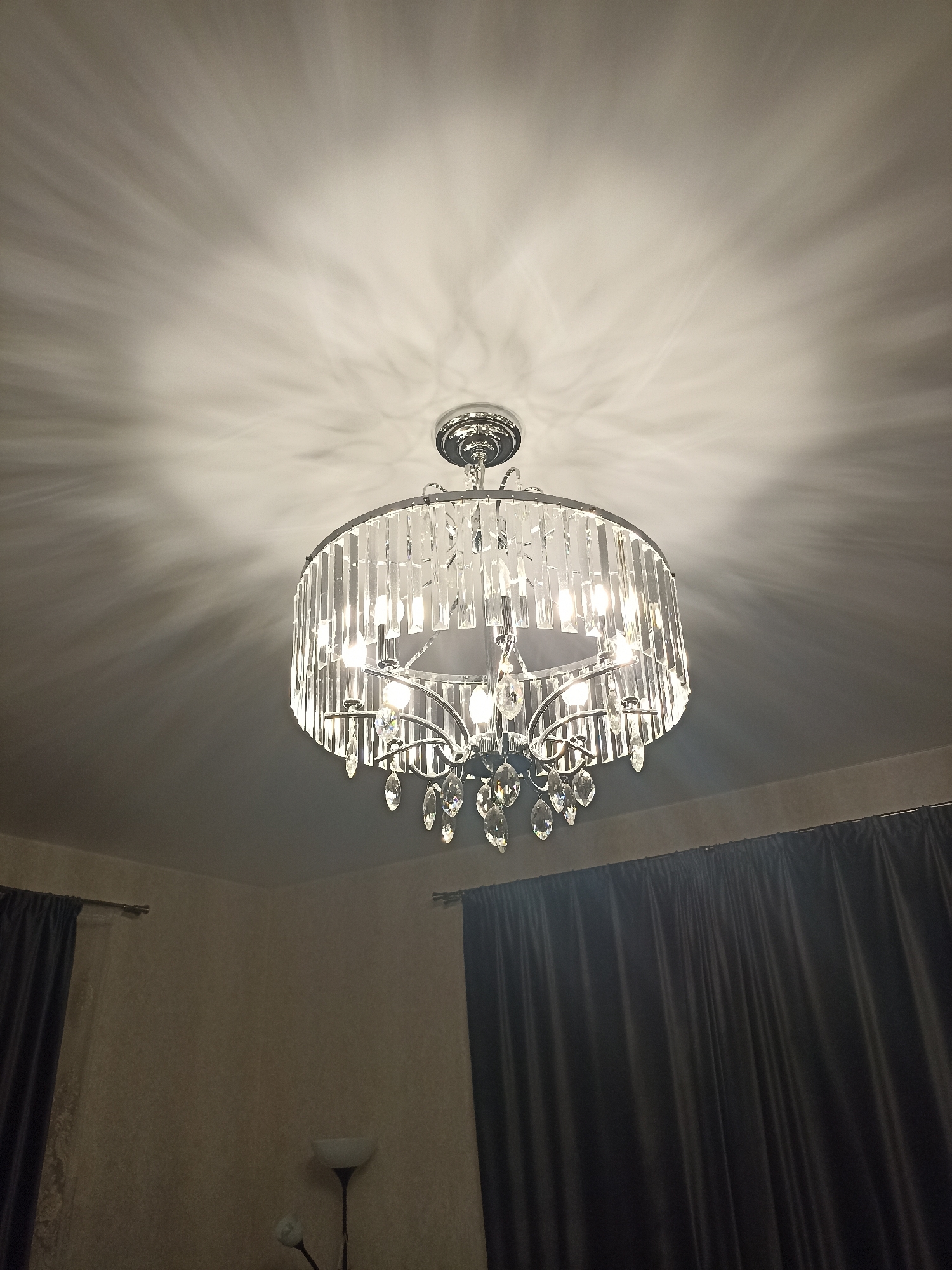 A contemporary crystal chandelier hangs from a white ceiling, casting intricate shadows across its surface. The chandelier is round and features multiple tiers of clear crystal prisms and teardrop-shaped pendants. Inside, several light bulbs illuminate the fixture. The ceiling is the primary focal point, with the rest of the room appearing dimly lit. In the lower portion of the image, dark gray curtains are visible on the right and left sides, framing the space. A floor lamp with a white shade stands in the lower left corner, partially obscured. The overall impression is of an indoor setting, likely a living room or dining area.