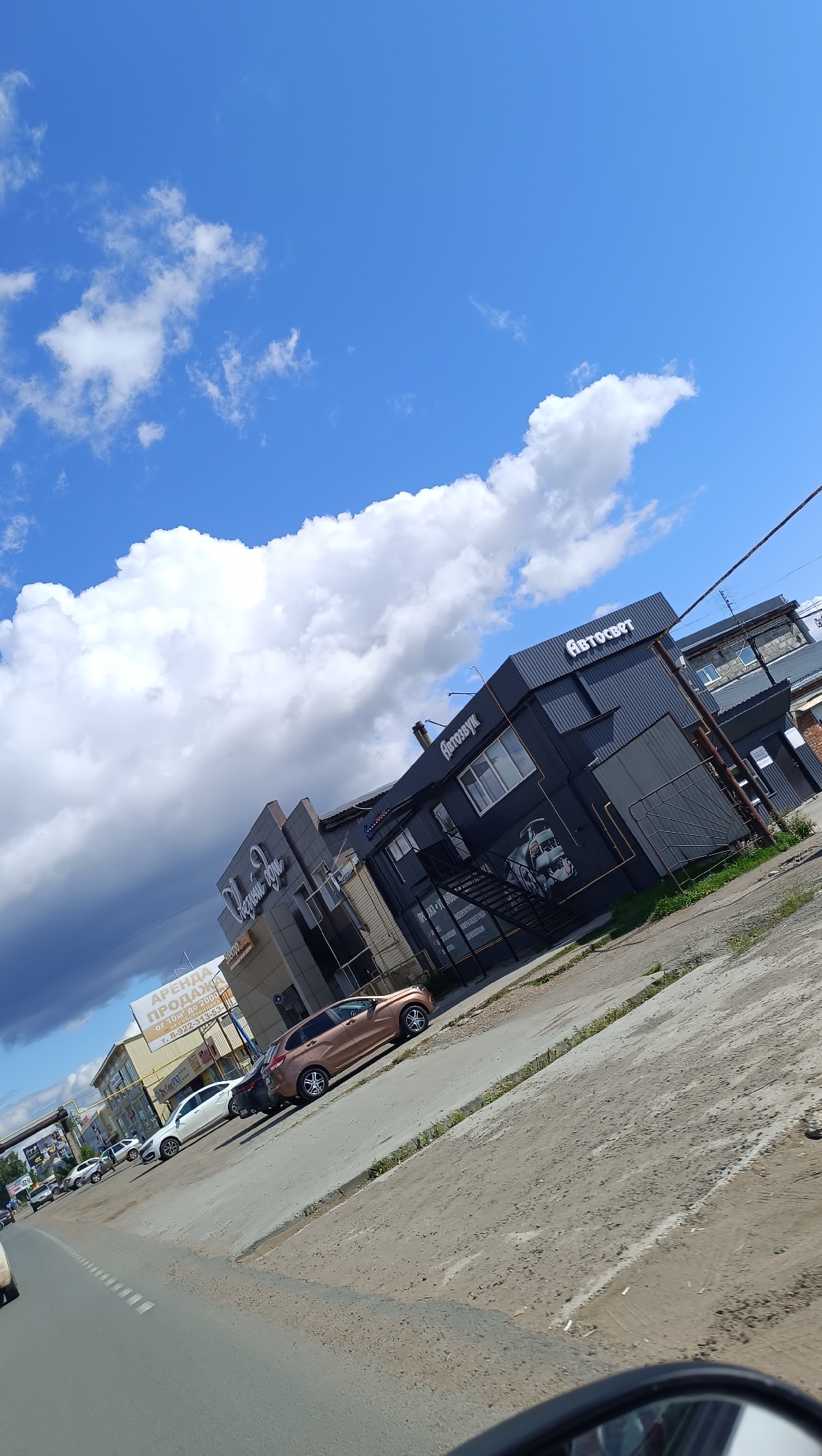 A vertical, slightly tilted, eye-level, outdoor, daytime shot captures a street scene in Chaikovsky, Russia. A bright blue sky with scattered, fluffy white clouds and a large, dark storm cloud dominates the upper portion of the image. In the mid-ground, a row of businesses with dark, corrugated metal facades lines the street. One building prominently displays the white text "АВТОЗВУК" (Autozvuk, meaning Car Audio) and a graphic of a car engine. Another building to its left has signage that includes "Аренда" (Rent) and "Продажа" (Sale). Several cars, including a light brown hatchback and a white sedan, are parked along the curb or on the street. The foreground shows a paved road with a dirt shoulder and some greenery. The perspective suggests the photo was taken from a vehicle.