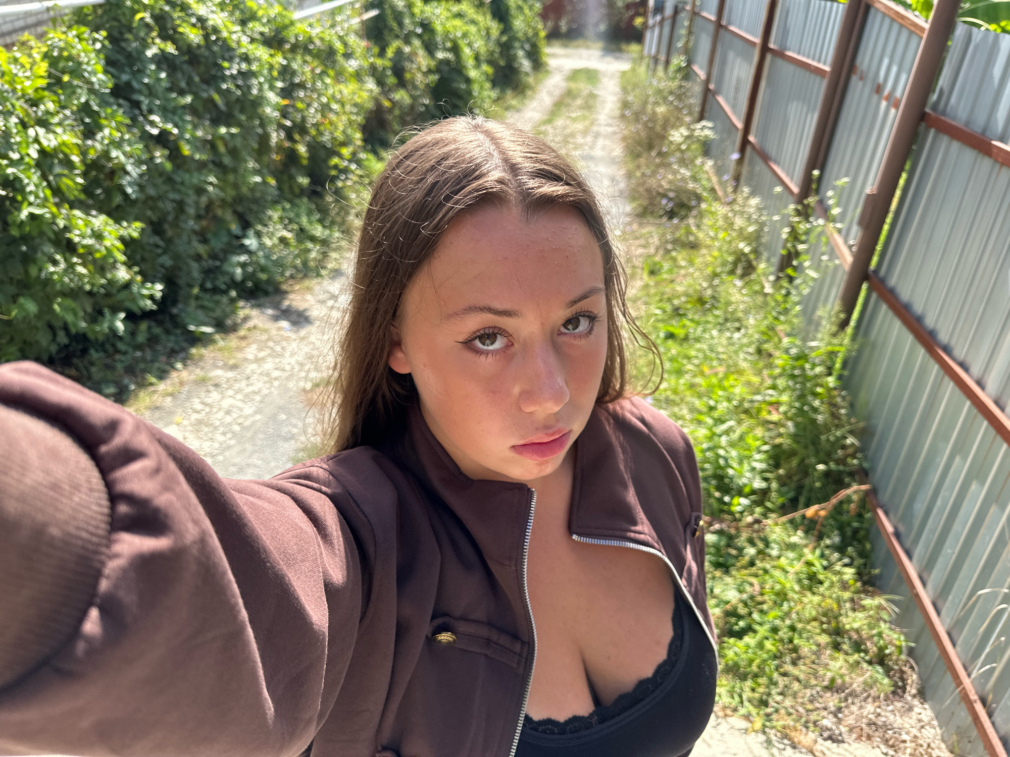 A young woman is taking a selfie in an outdoor setting, with a gravel path extending into the background. She is wearing a brown zip-up jacket over a black lace-trimmed top. To her left, lush green bushes line the path. To her right, a corrugated metal fence with wooden framing runs alongside the path, with wild grass and small plants growing near its base. The scene appears to be in Lermontovo, Russia.