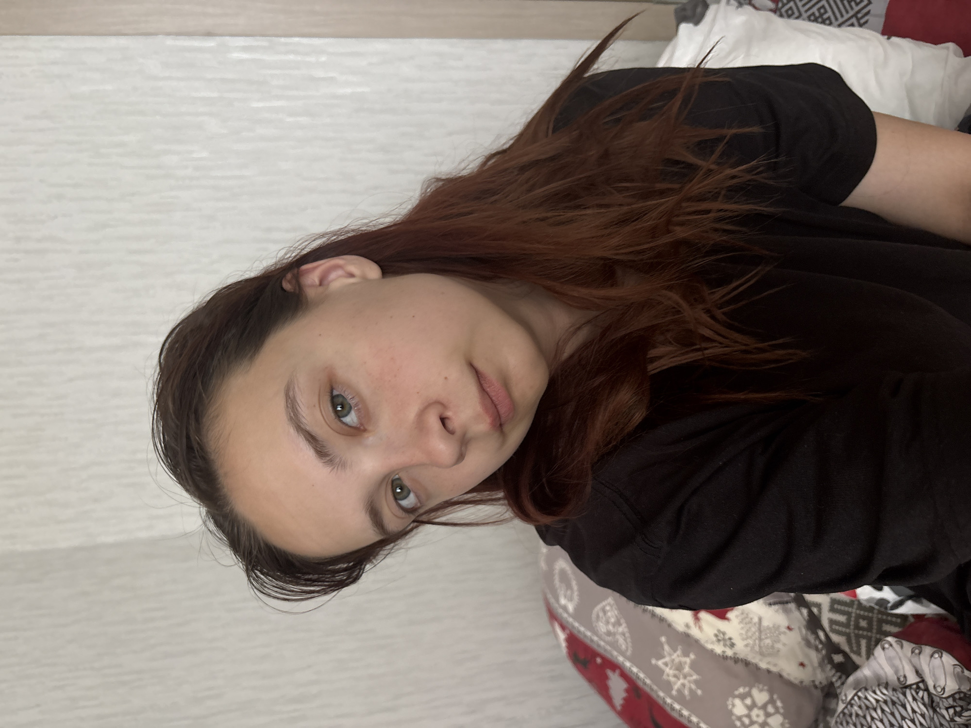 A close-up shot shows a woman with light skin, green eyes, and brown hair wearing a black shirt. The background features a wall with a light, textured pattern, and a patterned blanket is visible beneath her. The image's location is unknown and has not been given, nor could be inferred from the visual evidence.