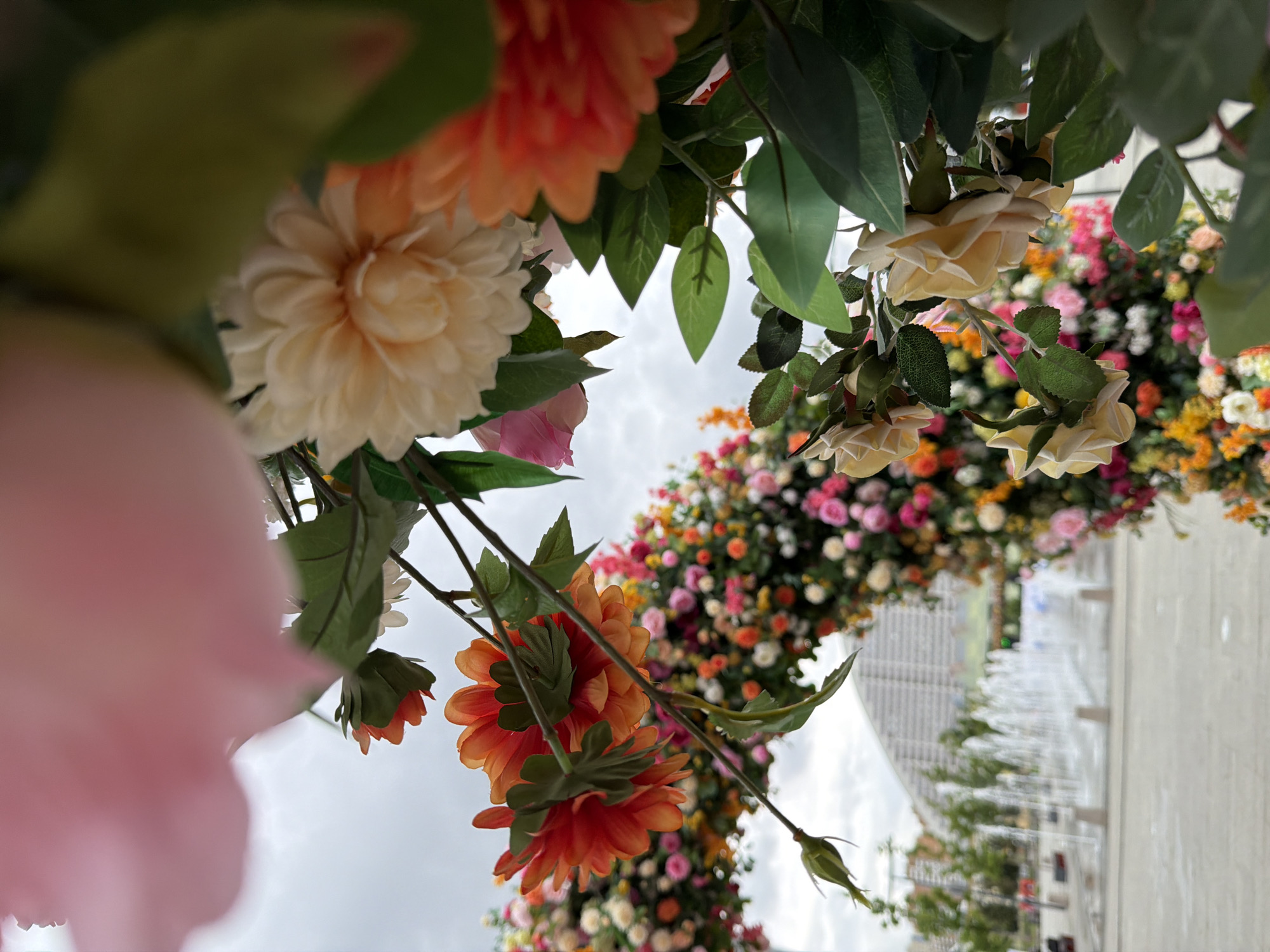This is a close-up, low-angle shot of a floral arrangement featuring a variety of artificial flowers in shades of peach, cream, yellow, and pink, along with green leaves. The flowers are densely packed, forming a decorative arch or display. In the background, a blurred scene depicts a modern building with a distinctive gridded facade, a water fountain feature with multiple jets spraying water, and some trees. The sky is overcast, suggesting a cloudy day. The foreground is dominated by the vibrant, textured petals of the flowers, with some elements, like a large pink petal on the left, being out of focus and very close to the camera. The perspective creates a sense of being immersed within the floral display. There is no visible text or identifiable people in the image. The scene appears to be outdoors in a public space, possibly a park or plaza, given the presence of the fountain and modern architecture. The overall impression is one of decorative floral artistry set against a contemporary urban backdrop.