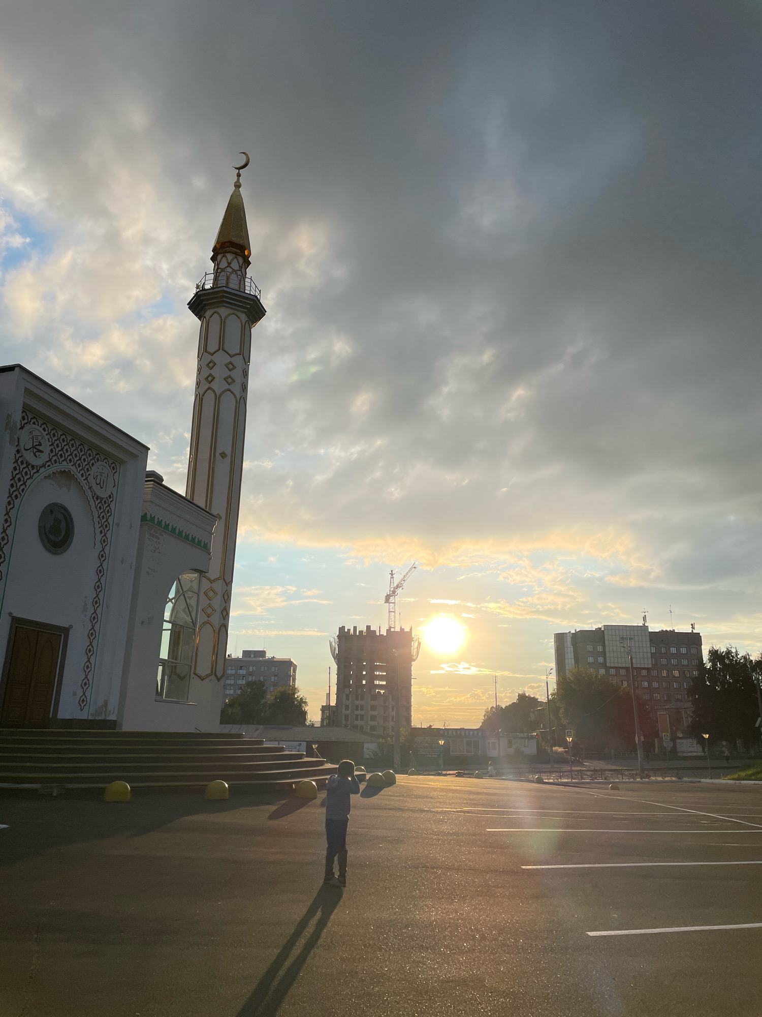 The image shows a mosque with a minaret, located in the city of Izhevsk, Russia. The mosque is white with gold trim and has a crescent moon on top of its minaret. In front of the mosque, there is a person standing and looking at the sun, which is setting in the background. The sun is casting long shadows across the paved area in front of the mosque. In the background, there are several buildings, including a construction site with a crane. The sky is cloudy, with the sun peeking through the clouds. The overall scene is one of a peaceful evening in a city.