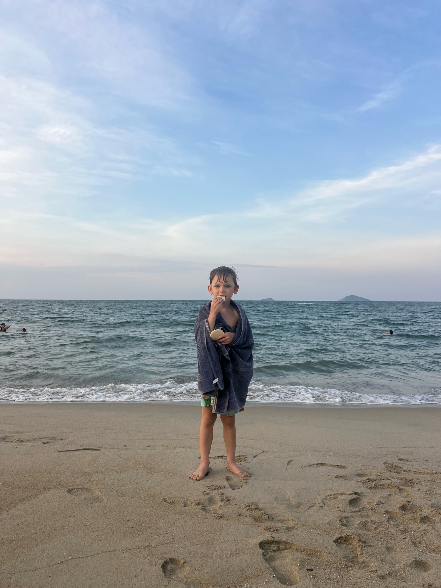 The image depicts a young boy standing on a sandy beach in Hoi An, Vietnam, with the ocean in the background. The boy is wrapped in a dark blue towel and is eating something light-colored. He is wearing patterned shorts. The water is calm with small waves, and there are other people visible in the ocean in the distance. The sky is partly cloudy with a light blue hue. Footprints are visible in the sand around the boy.