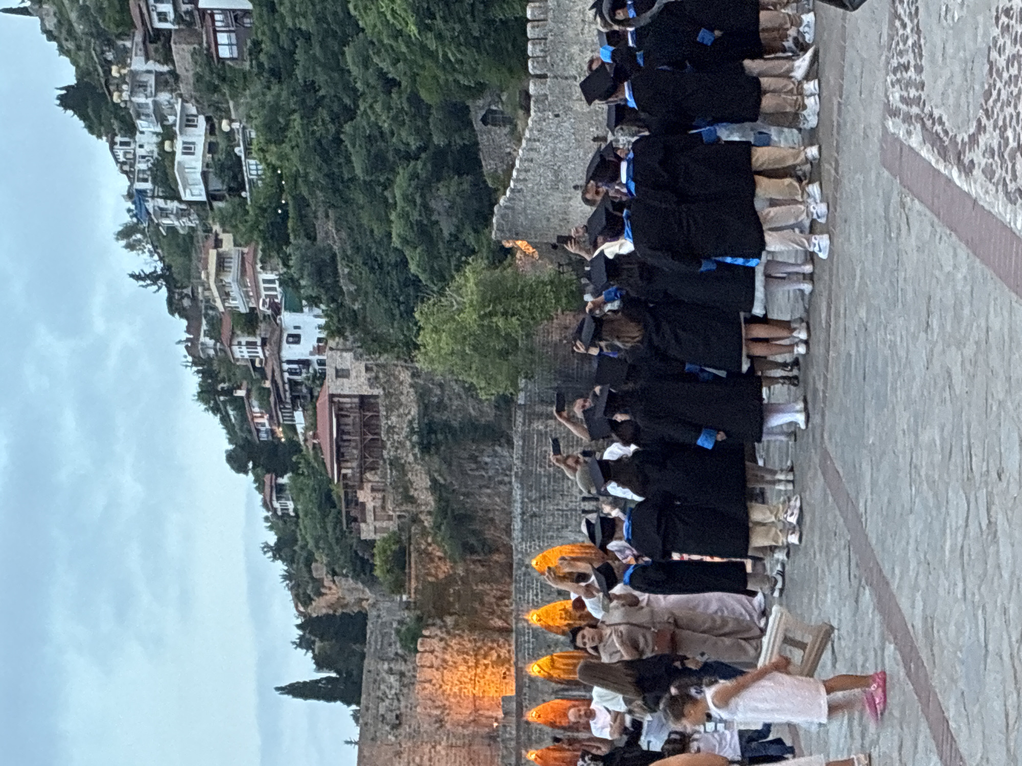 This media file captures an outdoor event, likely a graduation ceremony, taking place at dusk or on an overcast day in Alanya, Turkey, against the backdrop of a historic fortress, possibly Alanya Castle. The central subjects are a line of people, predominantly young adults, dressed in black graduation gowns and mortarboard caps, many featuring blue accents. Several individuals in the line are holding up smartphones, seemingly recording or photographing the event.
They are standing on a paved surface next to a sturdy, weathered stone wall with battlements. Beyond this wall, a dramatic landscape unfolds, featuring a steep, tree-covered cliffside dotted with numerous buildings, giving a sense of the town's mountainous setting. The sky is light gray and overcast.
In the immediate foreground, additional people are present, including adults sitting on chairs and a young child in a white dress and pink shoes. Several glowing orange or yellow objects, possibly lanterns or decorative lights, are arranged on the ground, contributing to the evening atmosphere. The scene evokes a celebratory and significant occasion set against a picturesque and historical backdrop. No visible text is discernible in the image.