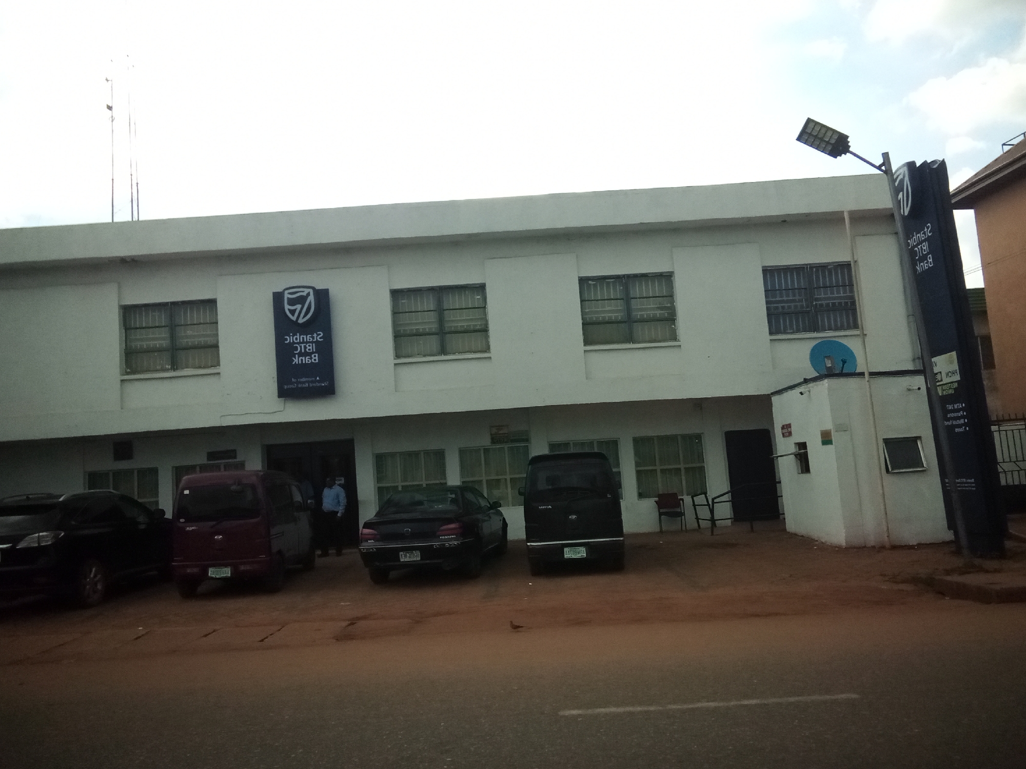 The image shows a street view of a Stanbic IBTC Bank branch in Nibo, Nigeria. The bank is a white, single-story building with several windows and an entrance. A tall, dark blue sign with the Stanbic IBTC logo and name is prominently displayed in front of the building. Several vehicles are parked in the lot in front of the bank, including a maroon minivan, a black sedan, and a black van. A person in a light blue shirt and dark pants is standing near the entrance. The area appears to be a commercial or business district. The sky is overcast, and the lighting suggests it is daytime.