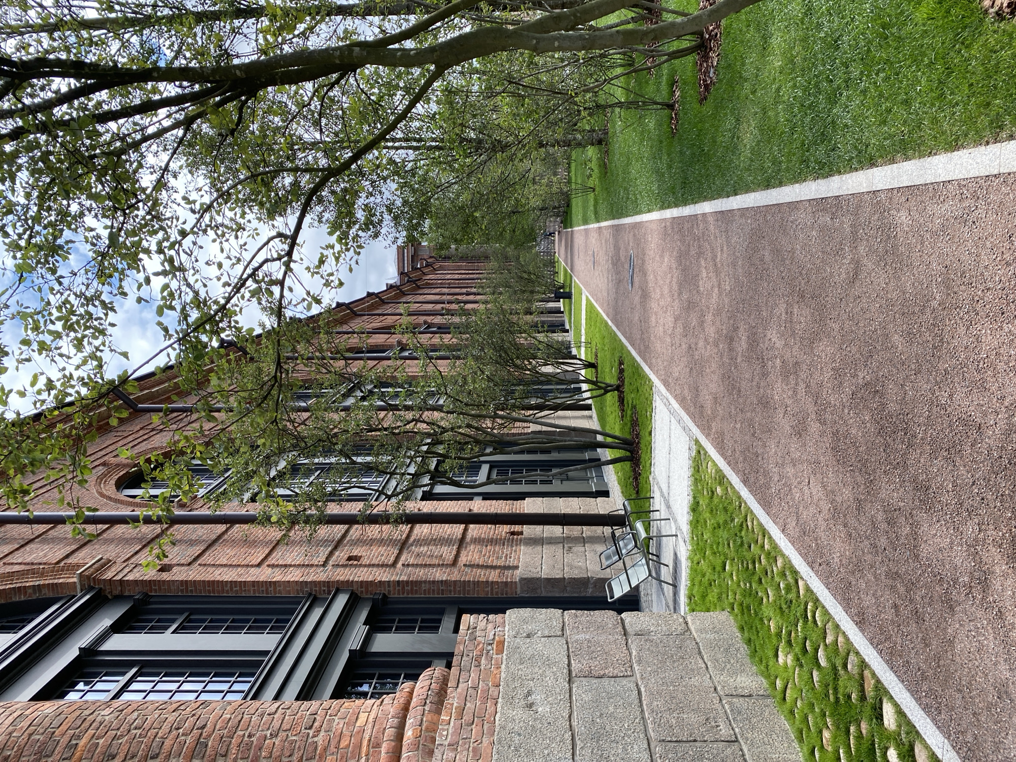 The image shows a view looking down a paved walkway in front of a long, red brick building with dark framed windows. The walkway is bordered by a strip of green grass on the right, and on the left, there is grass with round stones embedded in it, adjacent to the building's stone base. Two light-colored chairs are placed on the grass near the building. Several young trees with green leaves are planted along the walkway, mostly on the right side. The sky above is partly cloudy. This scene appears to be located at the New Holland Island complex in Saint Petersburg, Russia, known for its renovated industrial buildings and landscaped grounds.