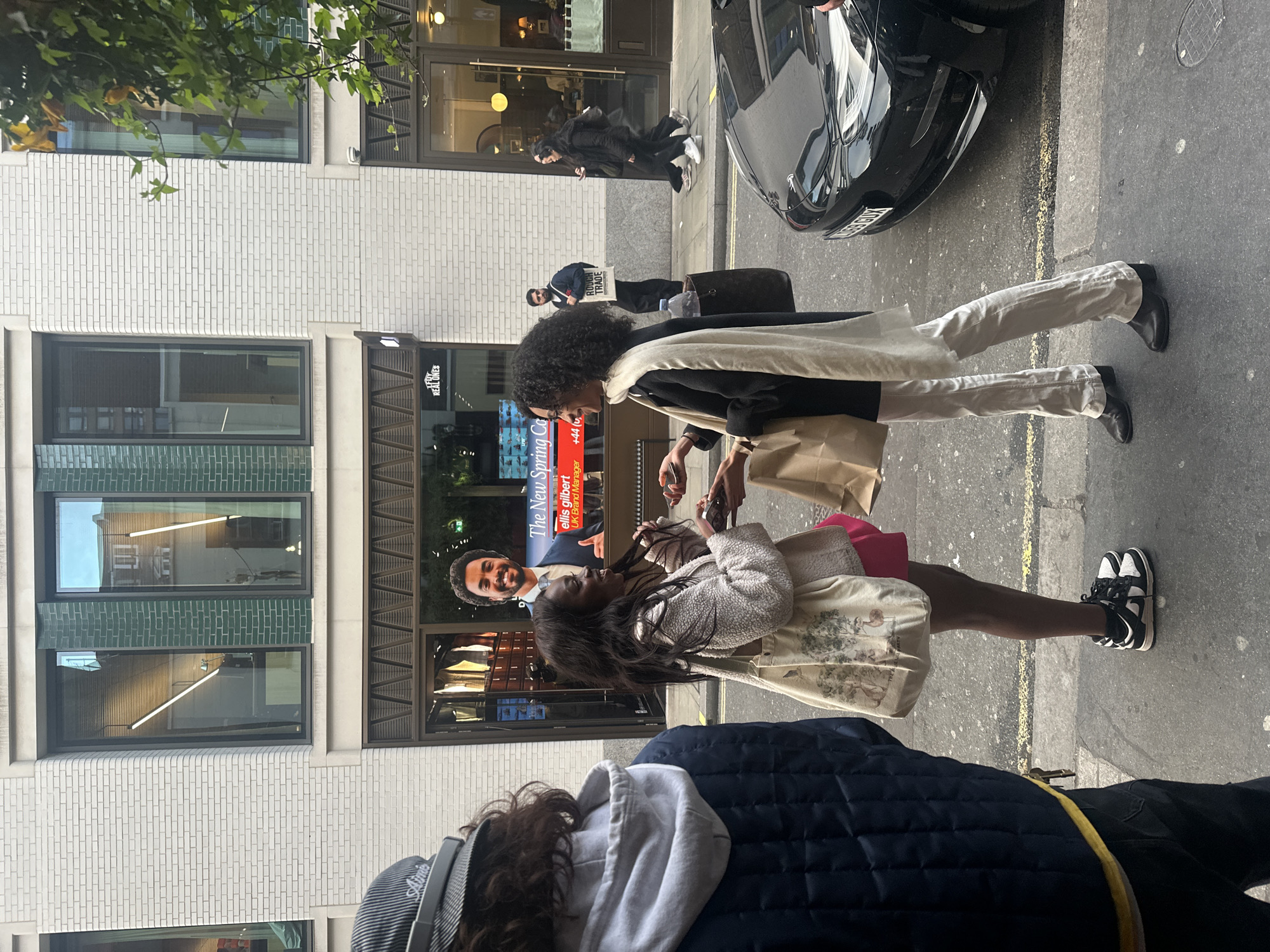 Here is a detailed description of the image:
**Scene Overview:**
The image captures a street scene in the city of London, United Kingdom.
**Foreground:**
The lower portion of the frame contains a person wearing a dark blue quilted jacket with a gray hoodie and a striped baseball cap. The person's face is not fully visible.
Two women are standing and interacting in the center of the image. One is wearing a black coat, light-colored trousers, and black boots. She has a white scarf draped around her neck and is holding a beige shopping bag. The second woman is wearing a cream-colored jacket, a light-colored skirt with illustrations on it, black and white sneakers, and carrying a pink bag.
**Background:**
The backdrop consists of buildings and the street. A black vehicle, partially visible, is parked on the side of the road. Other people are walking in the background, including one person holding a white sign with the words "ROUGH TRADE" visible.
A store with a window display featuring "The New Spring Co." advertisement and a portrait of a man named "Ellis Gilbert, UK Brand Manager," along with a phone number and the text "REAL ONES" is visible.