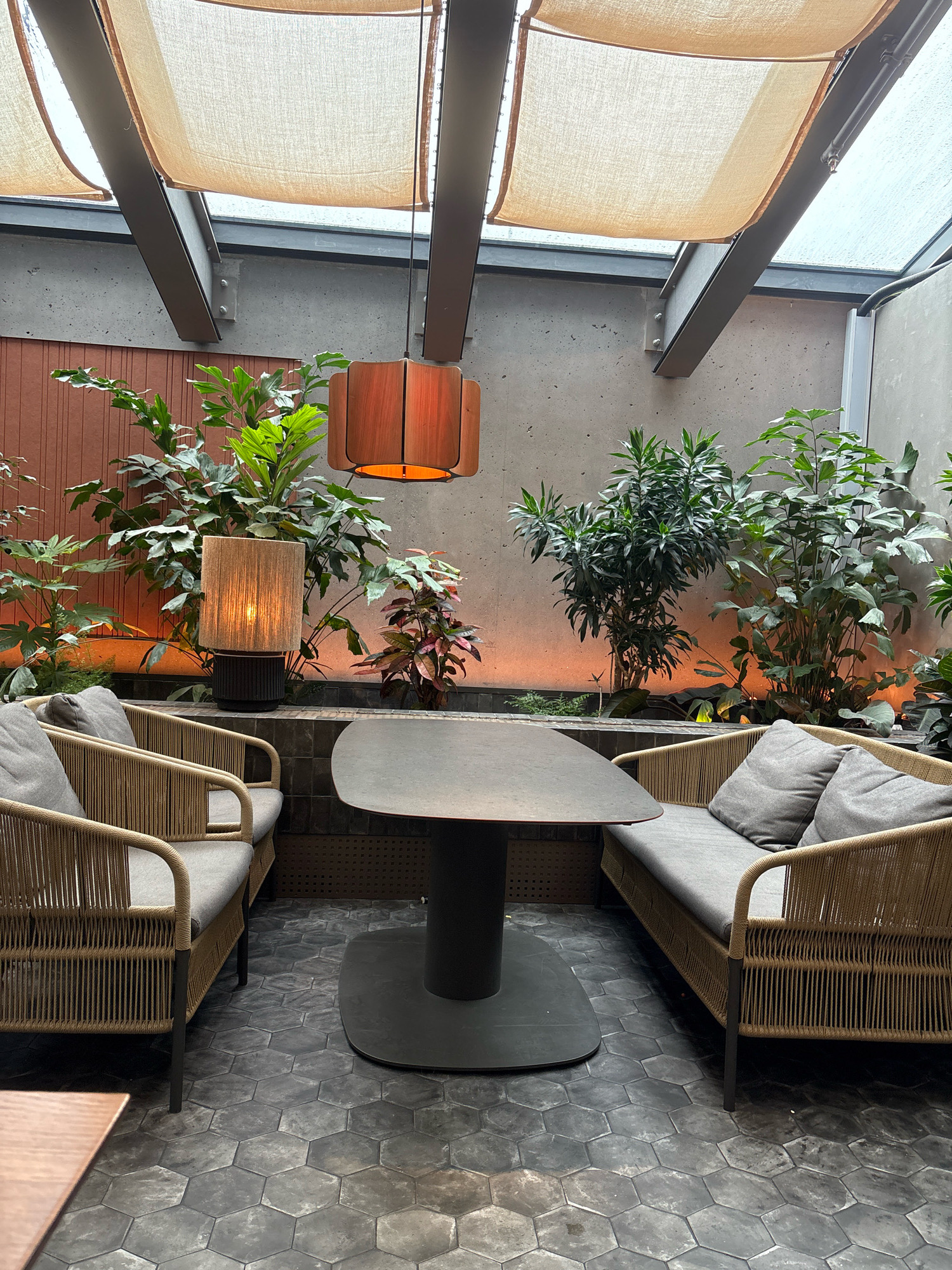 This is a medium shot, eye-level, indoor photograph of a seating area with lush greenery, located in Kopavogur, Iceland. The scene is dimly lit, with warm ambient light emanating from two lamps. On the left, a woven armchair with grey cushions is visible. To the right, a woven loveseat with grey cushions is positioned opposite the armchair. Between them is a dark, oval-shaped table with a pedestal base. The floor is covered in dark, hexagonal tiles. Behind the seating arrangement, a variety of plants are situated in planters. The background features a textured brown wall on the left and a concrete wall on the right. Above, a ceiling structure is partially covered by beige fabric awnings, with exposed metal beams. A large, wooden pendant lamp hangs from the ceiling. The overall impression is that of a cozy, naturalistic indoor space.
