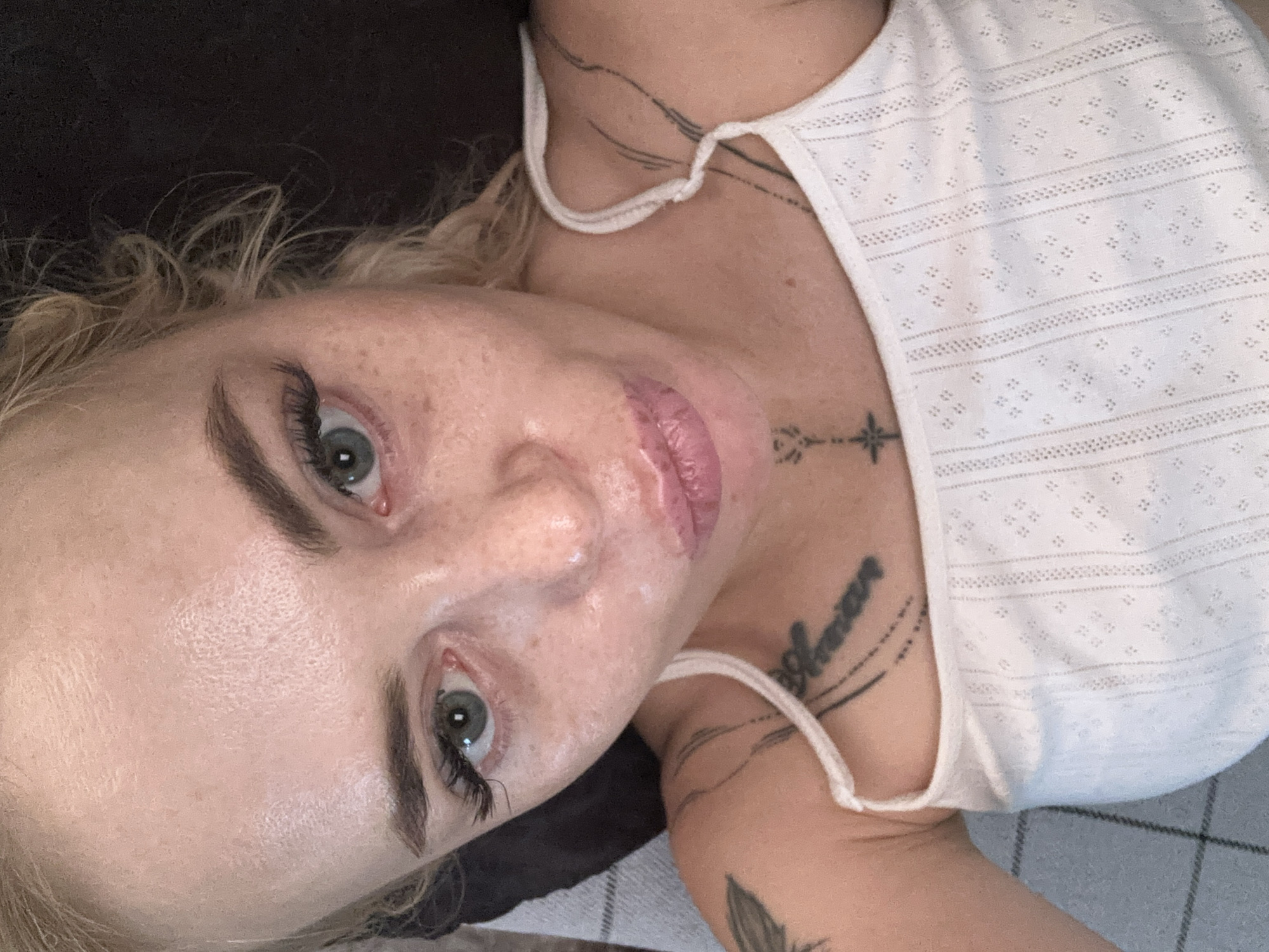 A young woman with blonde hair and blue eyes is shown in a close-up, overhead shot. She is wearing a white tank top and has a tattoo on her left shoulder. Her lips are slightly parted, and she appears to be looking directly at the camera. The background is dark and out of focus. The lighting is soft and natural.