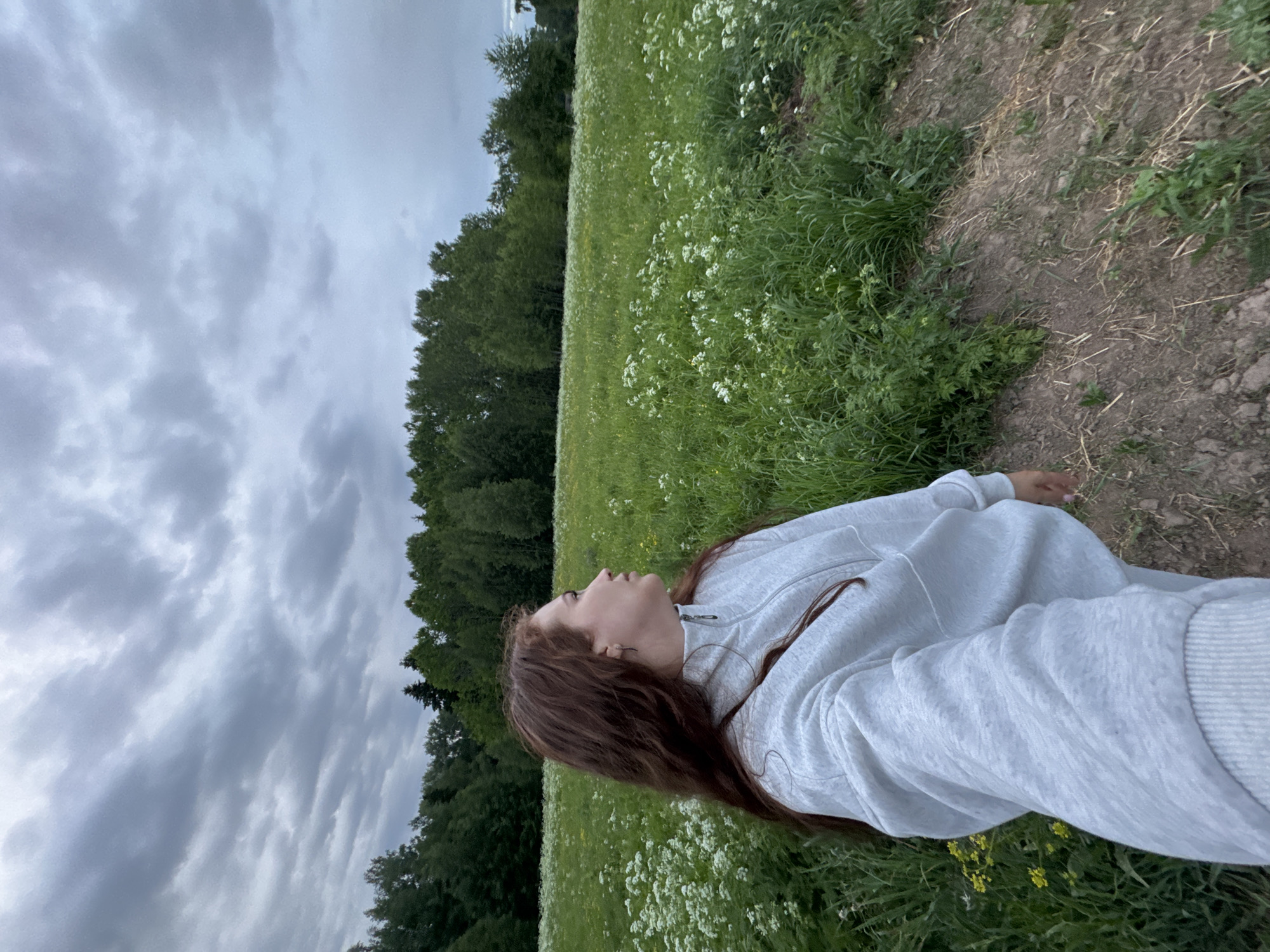 A woman with long, reddish-brown hair lies in a grassy field with scattered white wildflowers, possibly cow parsley, and some yellow flowers. She wears a light gray hooded sweatshirt and appears to be looking up at the sky. Behind her is a line of trees, and the sky is filled with gray clouds, suggesting an overcast day. The ground near the edge of the field is dirt or bare earth. Location is likely near Nizhnyaya Gar', Russia.