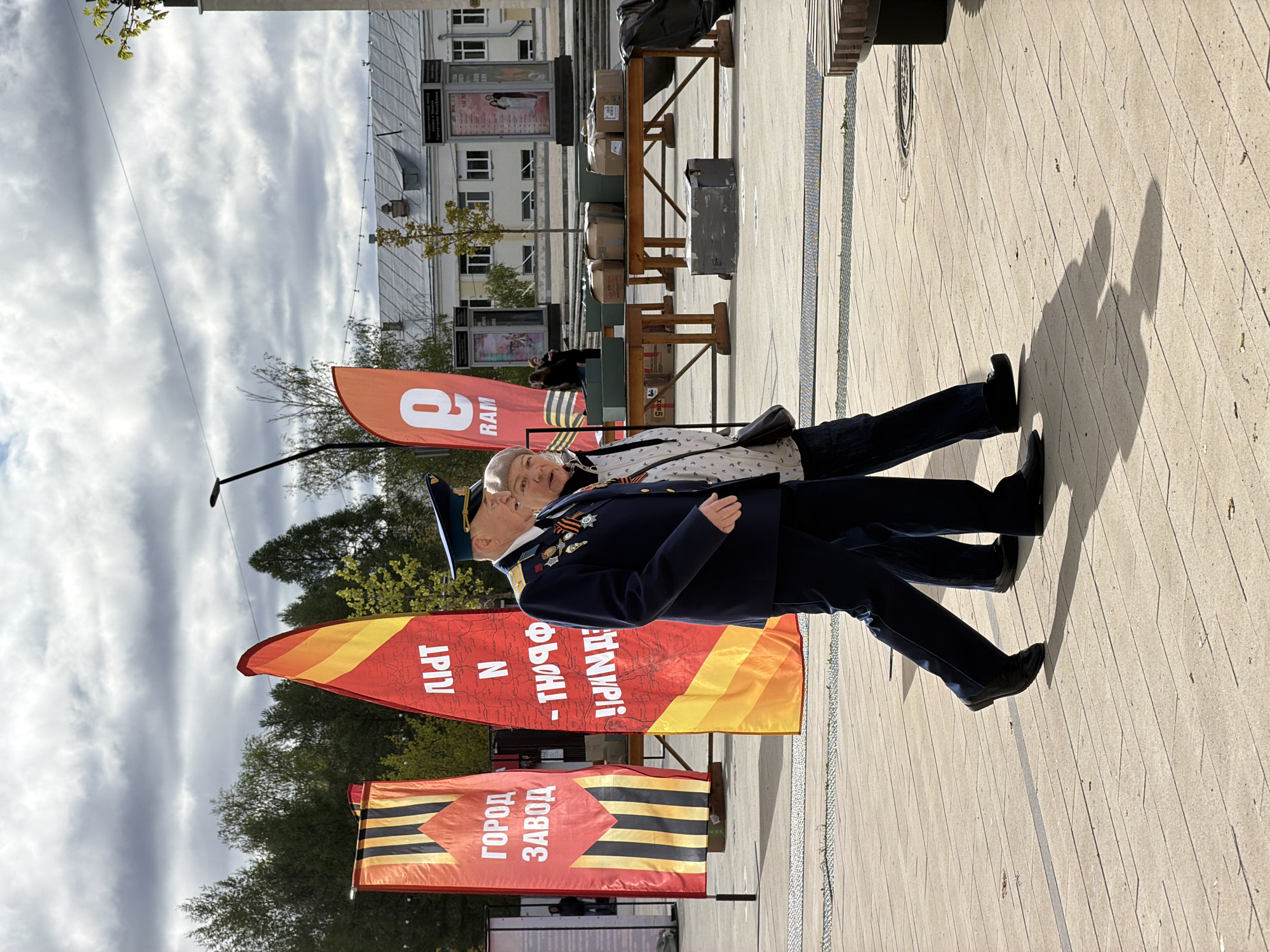 In Izhevsk, Russia, a man in a dark blue military-style uniform stands with a woman on a paved surface. The man wears a peaked cap and several medals are visible on his chest. Two large, colorful flags are visible; one reads "ГОРОД ЗАВОД" (City Factory) and the other includes the word "ИЖДЕРПРИМ." Buildings are present in the background, beneath a sky with scattered clouds.
