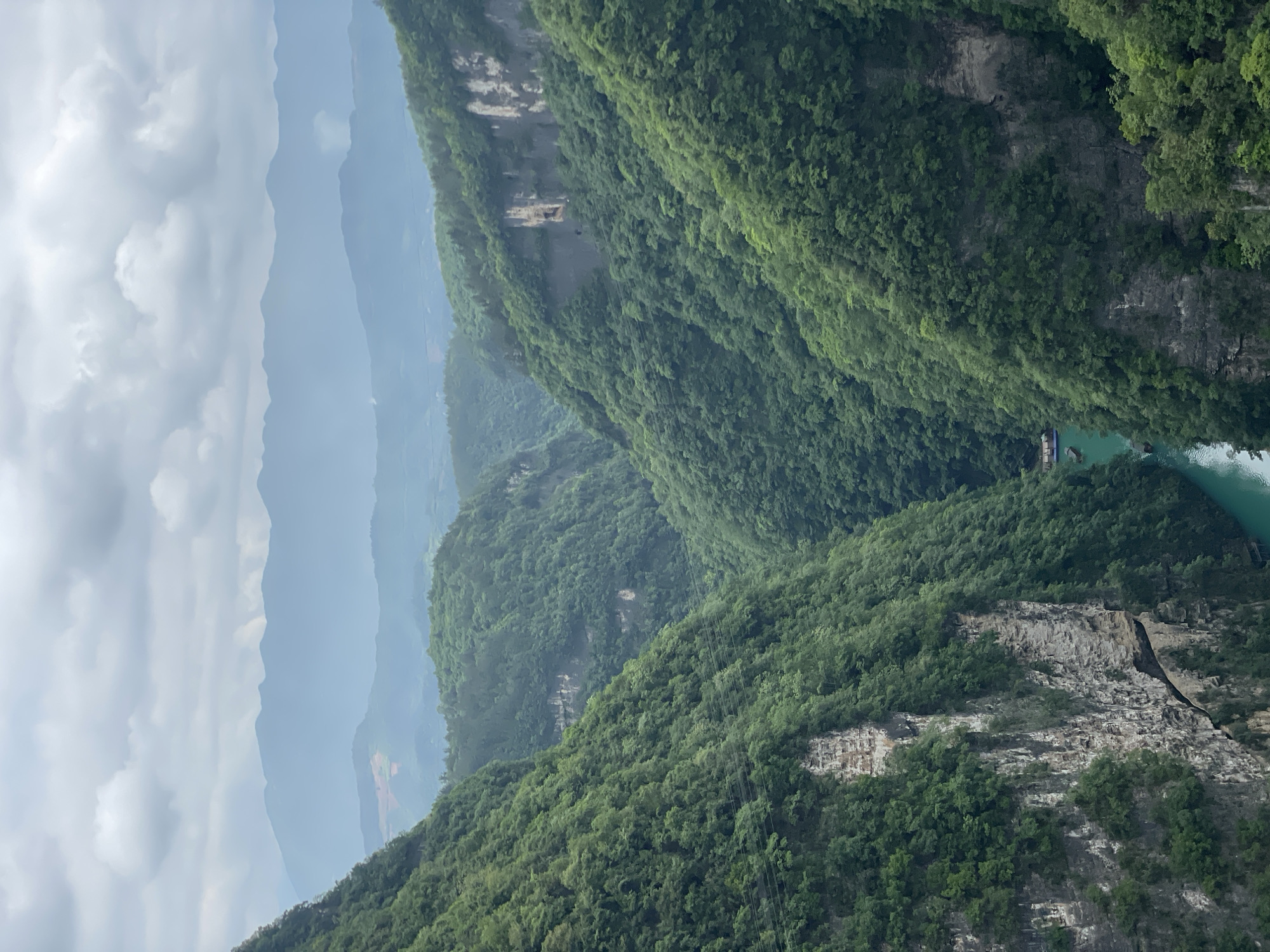 The image shows a view of forested mountains and a river in Zhangjiajie, China. The mountains are steep, covered in dense green foliage, and interspersed with rocky outcroppings. In the distance, layers of mountains are visible, fading into the sky. A body of water is visible between the mountains. The sky is overcast.