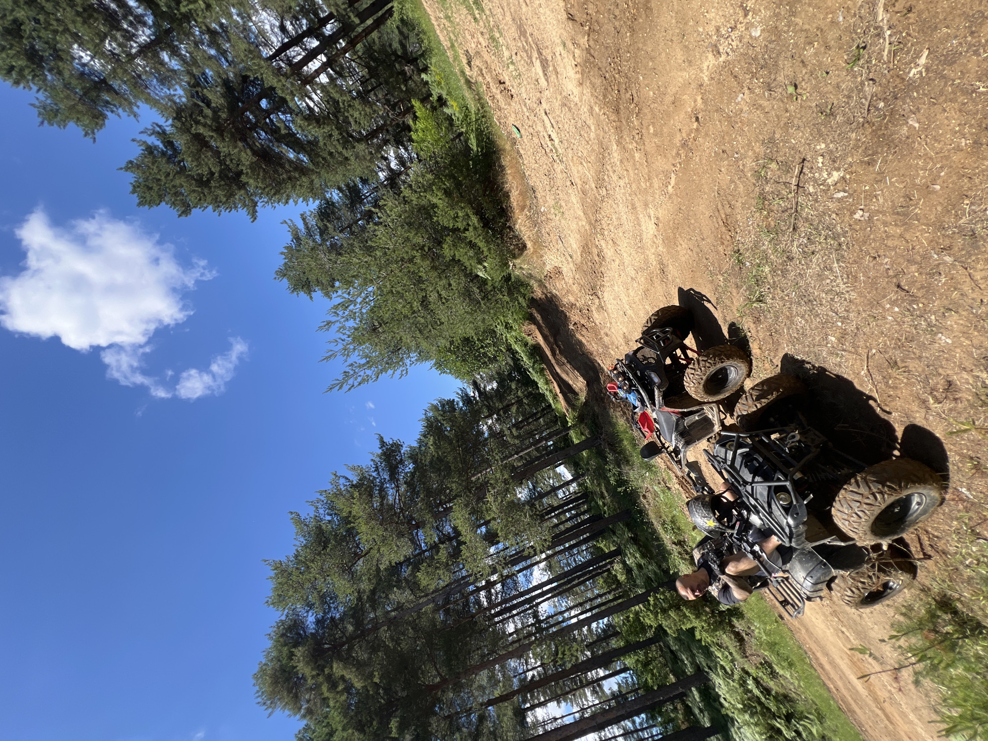 The image shows a sunny day in a wooded area, likely Djerovo, Bulgaria. A man is seated on a black ATV, wearing a black t-shirt and a helmet. Another red ATV is parked behind him. The ATVs are on a dirt path surrounded by tall pine trees and green foliage. The sky is blue with a few white clouds. The scene suggests an outdoor recreational activity, possibly off-roading.