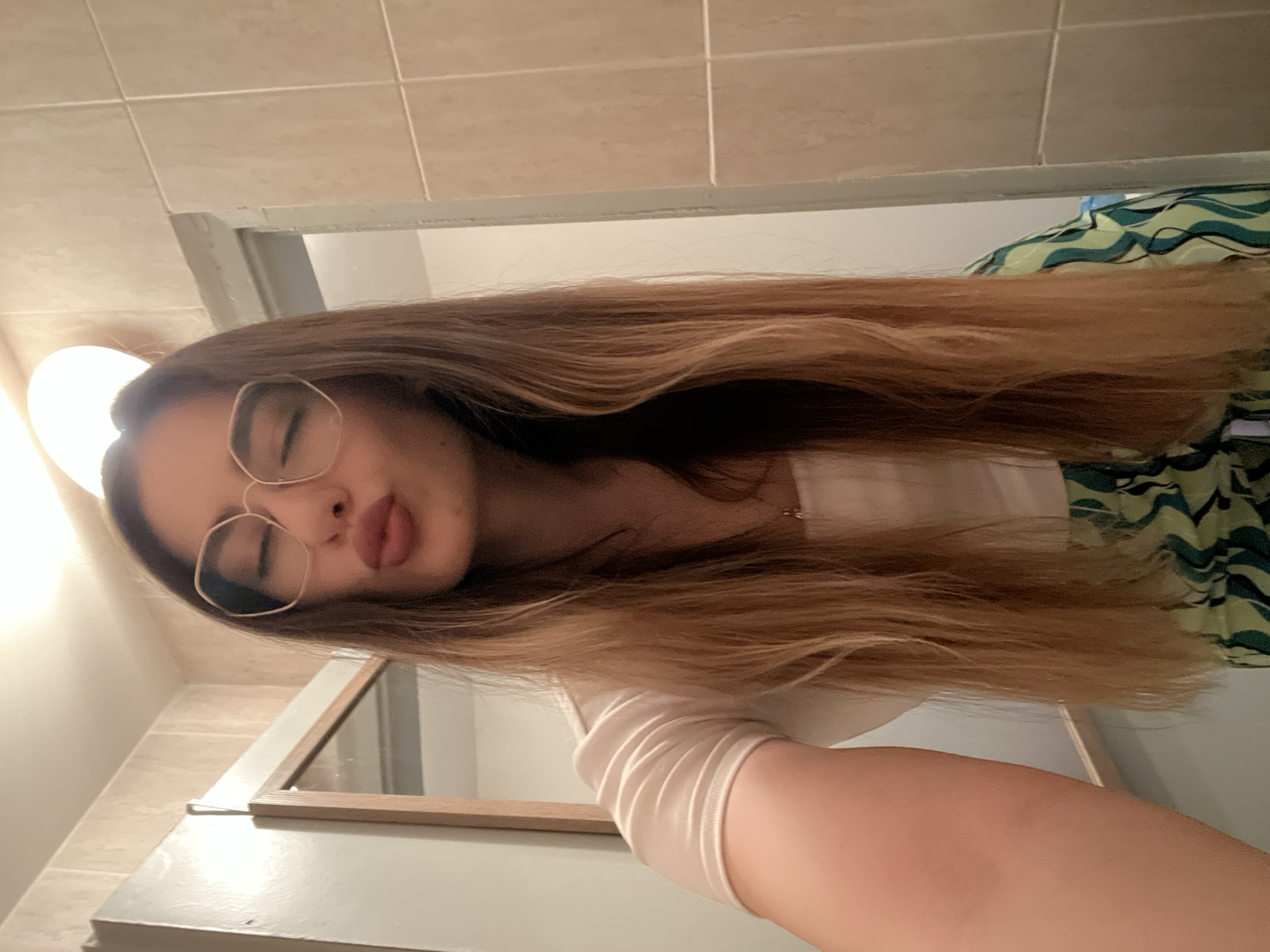 A selfie portrait of a young woman with long, light brown hair and glasses. She is wearing a light-colored top and has her eyes closed and lips pursed. The background appears to be a bathroom setting with light-colored tiles and a mirror. A green and white patterned object is visible in the upper right corner. The location is Unknown, Unknown.