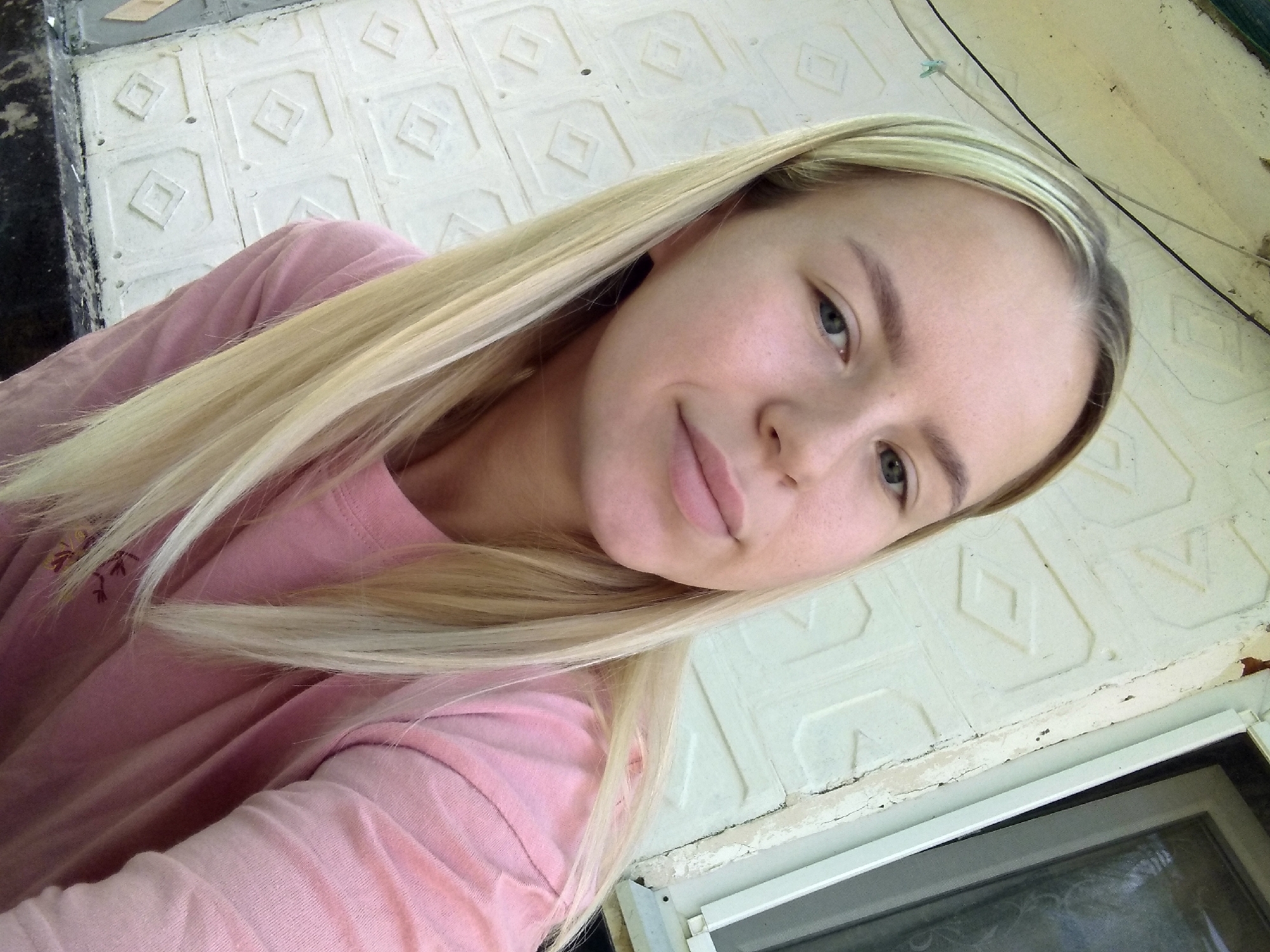 A young woman with blonde hair is pictured outdoors, with a white textured wall and a window visible behind her. She is wearing a pink long-sleeved shirt. The wall has a repetitive diamond pattern. There is a wire running across the top of the image. The scene appears to be in a residential area.
