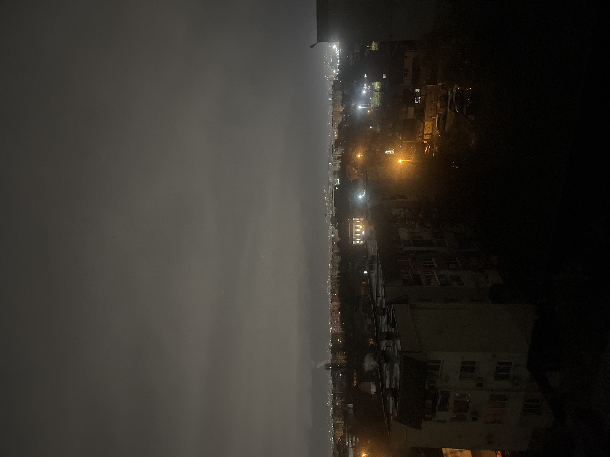 The image shows a night view of Sofia, Bulgaria from an elevated position, likely a balcony. The city lights stretch into the distance, partially obscured by fog or smog. To the right, dark buildings are visible, while to the left, the sky is dark and overcast. There are no visible people or interactions in the frame.