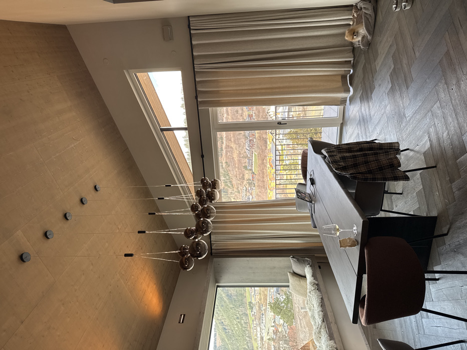 The image depicts the interior of a modern dwelling, likely a dining area, offering a panoramic view of the city of Schladming, Austria, and its surrounding mountainous valley. The room features a high, sloping ceiling constructed from light-toned wooden planks. Seven individual pendant light fixtures, each comprising multiple smoked glass spherical globes suspended at varying heights, are mounted along the ceiling's slope.
On the left, a large rectangular window extends from floor to ceiling, revealing a vista of residential buildings with colored roofs nestled within the valley, surrounded by wooded hills. Below this window, a built-in window seat with light-colored cushions, a faux fur throw, and two accent pillows is present. Adjacent to the window, a glass door opens to an exterior balcony, with a metal railing visible. Above this door, a triangular window allows additional light. Floor-length, light beige curtains are drawn back, framing the large window and glass door.
A large, dark-colored wooden dining table with an irregular edge dominates the center of the room. Six modern dining chairs surround the table: two are upholstered in dark brown, two in light grey, and two in dark grey. A plaid shirt in brown, black, and white is draped over the back of one chair. On the table, an open laptop, a charging brick, and cable are situated at the far end. Closer to the foreground, a white wine glass containing a light-colored liquid and a small crumpled brown paper bag are visible.
The floor is laid with dark grey or dark brown wood in a herringbone pattern. In the lower right quadrant of the image, a small, light brown, fluffy dog rests within a soft grey pet bed. Two metallic dog bowls are placed on the floor near the dog bed. No people are present in the scene.