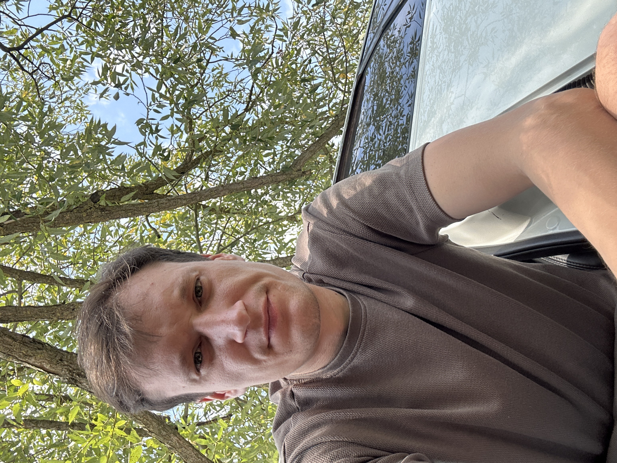 A medium shot photo shows a fair-skinned man with short, dark brown hair and light brown eyes. He is looking directly at the camera with a neutral expression, his mouth slightly open. The man is wearing a brown, short-sleeved t-shirt with a textured fabric. He is positioned in front of a white car, with the car's windshield and roof visible to his right. The background is dominated by green tree leaves and a bright blue sky. The overall lighting suggests it is daytime, possibly outdoors in a park or a natural setting. The image was taken in Makiv, Ukraine.