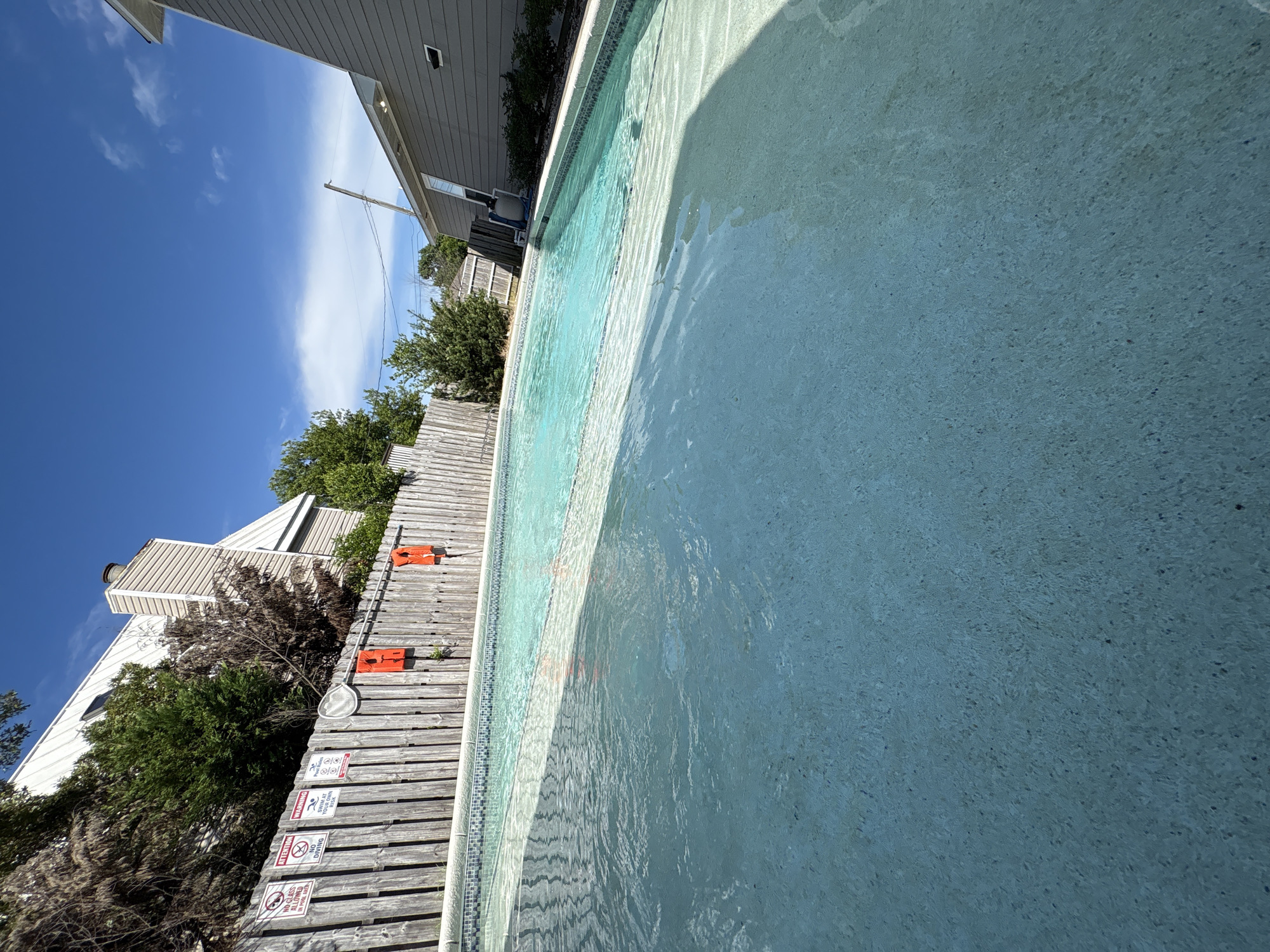 The image depicts a swimming pool in a residential setting, likely in Panama City Beach, Florida, given the clear blue sky with scattered clouds, lush green vegetation, and the style of the houses. The pool has a light blue-green water color and a textured, light-colored bottom. There are steps leading into the pool, indicated by a lighter shaded area. Along the edge of the pool, a tiled border is visible.
Behind the pool, a weathered wooden fence runs horizontally across the image. Attached to the fence are several warning signs and two bright orange life preservers. One life preserver is slightly to the left of the center of the image, and another is to the right. Above the fence, the roofs of houses and mature trees are visible. The house to the right has gray siding and a white trim. A utility pole with wires is visible in the background, extending towards the sky. The overall impression is that of a sunny day in a suburban or vacation home environment. There are no people or animals present in the image.