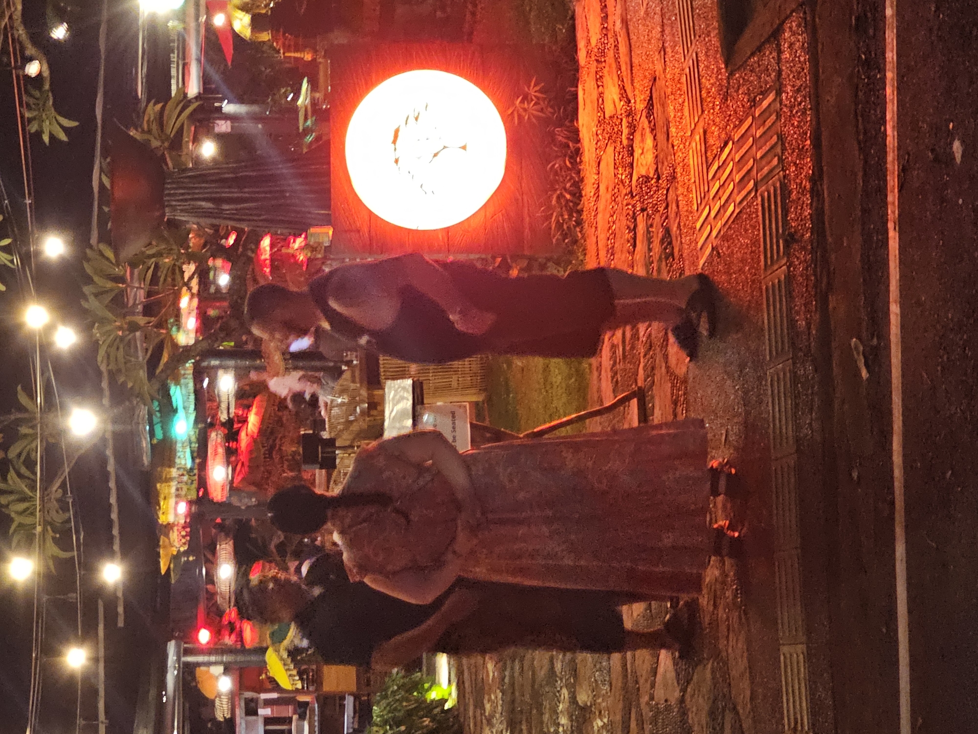 The image depicts an outdoor scene in Denpasar, Indonesia, at night. The location appears to be a restaurant or entertainment venue with a decorative, glowing red circle in the background. String lights illuminate the area. Three people are visible: one man in a dark tank top and shorts, and a woman in a floral dress. A third person is partially visible in front of the woman, wearing a dark shirt and holding a yellow object. All three are interacting, seemingly in conversation. The environment includes decorative lights, plants, and what appears to be outdoor seating. The ground is paved.