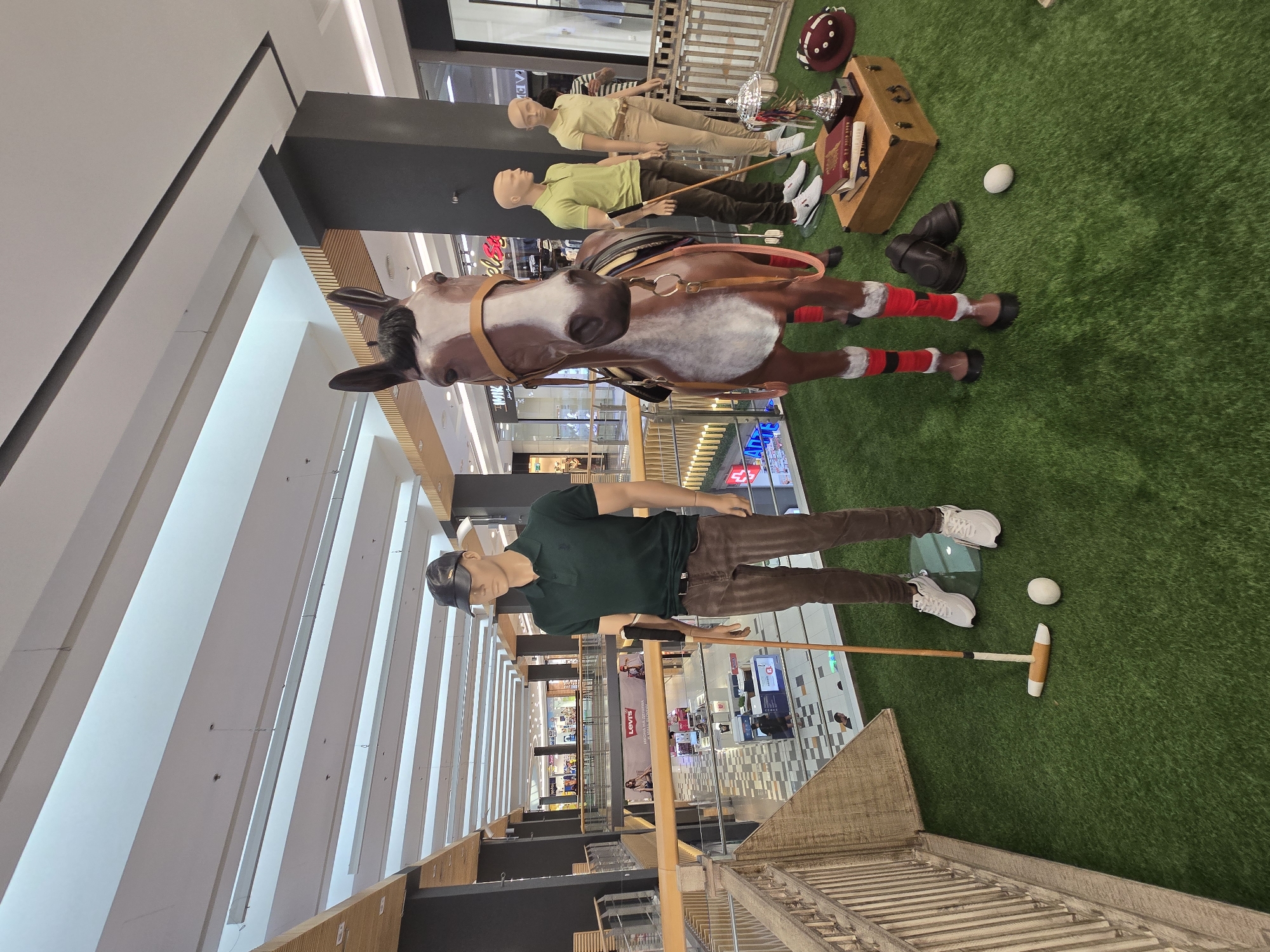 This image captures a store display inside a shopping mall, likely in Plovdiv, Bulgaria, given the Cyrillic text visible on some store signs in the background. The display features mannequins and a horse statue arranged on a green, artificial turf surface, simulating a polo or equestrian theme.
In the foreground, a male mannequin dressed in a dark green polo shirt, brown pants, and white sneakers stands with a polo mallet in his right hand, poised as if about to strike a white ball. A white polo ball rests on the turf near his feet. To the right of the mannequin, a large, brown and white horse statue is positioned. The horse is outfitted with a saddle, bridle, and red leg wraps.
Further to the right, two more mannequins are visible. One, dressed in a light green polo shirt and tan pants, holds a polo mallet. The other, in a light yellow polo shirt and tan pants, stands beside them. In front of these mannequins, a wooden chest serves as a base for a trophy, two books, and a dark red helmet with white polka dots. A pair of dark riding boots and another white polo ball are also on the turf near the horse and chest.
The background reveals the multi-level interior of a modern shopping mall with shops, escalators, and railings. The architecture includes light-colored ceiling beams and dark pillars. The overall scene is well-lit, characteristic of an indoor retail environment.