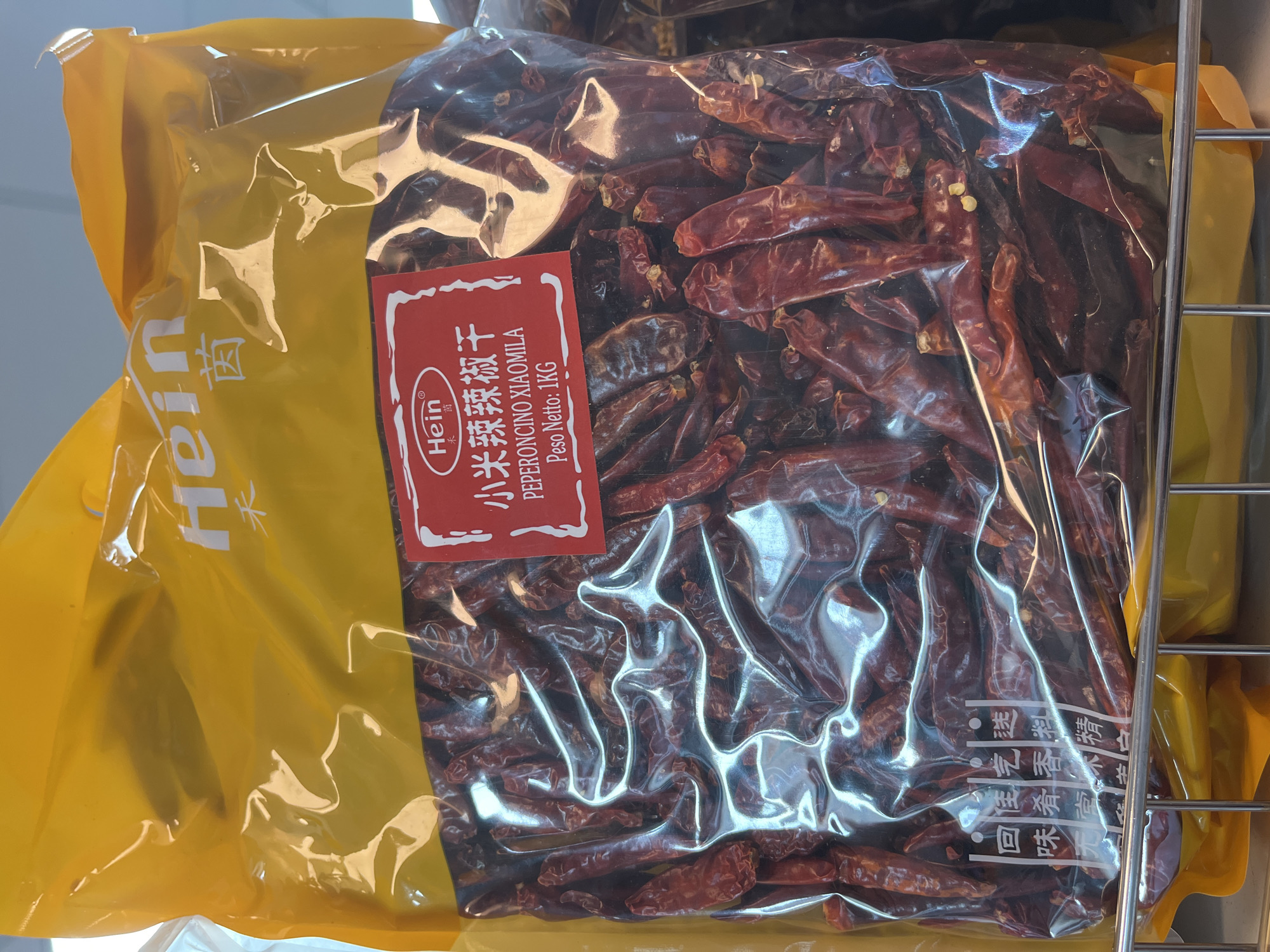 A large, clear plastic bag filled with dried red chilies is displayed on a metal shelf in what appears to be a grocery store or market. The bag is sealed with a yellow header that has white text, some of which is partially obscured by reflections. A red and white label is affixed to the front of the bag, featuring text in both Chinese and Italian. The Chinese text reads "小米辣辣椒干" and "Peperoncino Xiaomila", with "Peso Netto: 1KG" below it. In the bottom left corner of the bag, there is a vertical column of Chinese characters. The overall impression is that of a product for sale, specifically dried chili peppers.