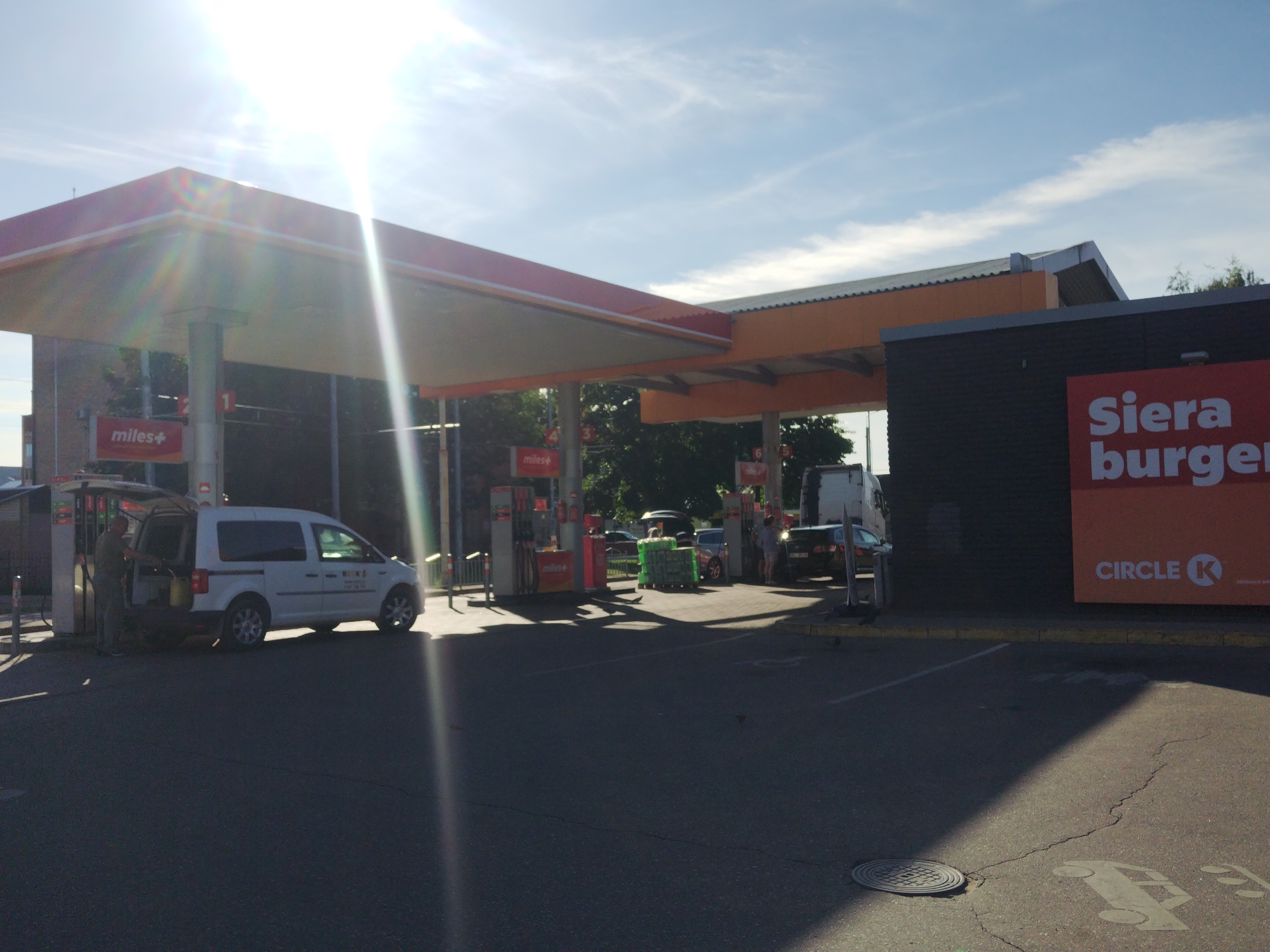 The image depicts a Circle K gas station and convenience store in Daugavpils, Latvia. The scene is set on a sunny day with bright sunlight causing lens flare in the upper left portion of the image. The gas station features a prominent red and orange canopy covering multiple fuel pumps. A white van with its rear hatch open is parked at one of the pumps, with a person standing beside it, possibly engaged in refueling or loading/unloading. Several other vehicles, including a black car and a white truck, are visible in the background near the store. The building itself has a dark facade with a large red sign displaying "Siera burger" and the Circle K logo. The foreground is an asphalt surface, likely a parking lot or roadway, with painted parking lines and a manhole cover. In the distance, trees and foliage can be seen, suggesting a semi-urban or suburban setting. The overall impression is that of a typical service station environment.